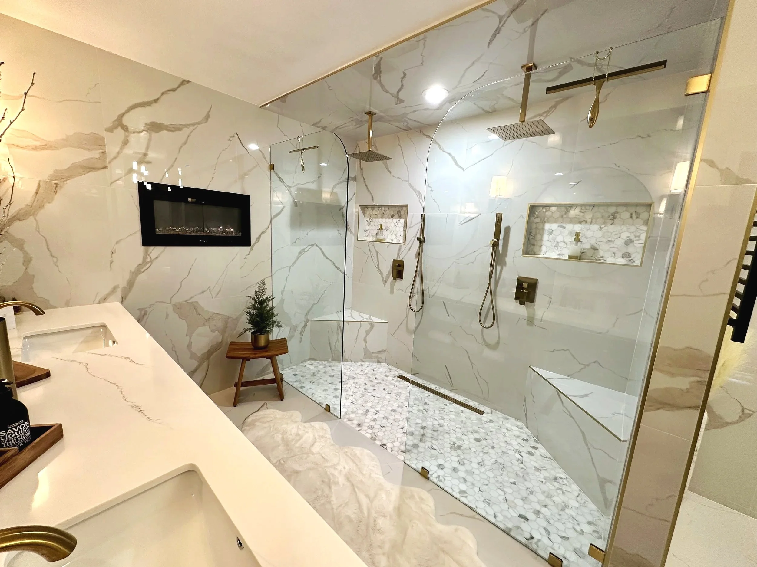 Modern bathroom with white marble walls and a large glass shower enclosure featuring two rain showerheads and textured pebble flooring. The shower has built-in niches with small bottles and a small potted plant on a wooden stool. There is a vanity with a white countertop and brass fixtures, and a wall-mounted fireplace adds a cozy touch.