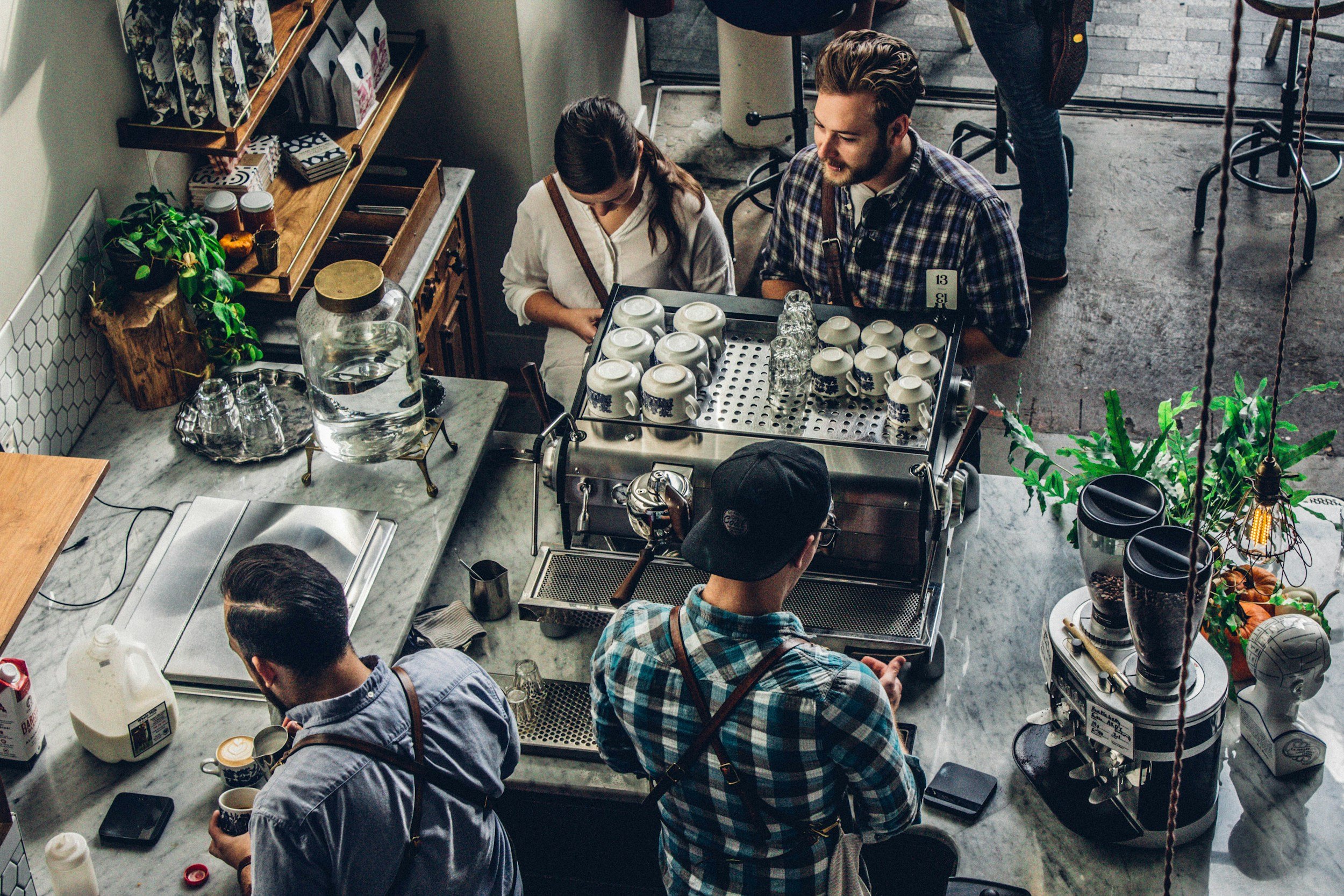 Overhead view of a busy coffee shop counter with baristas preparing drinks and customers ordering, with various coffee-making equipment, cups, and plants on the marble countertops.