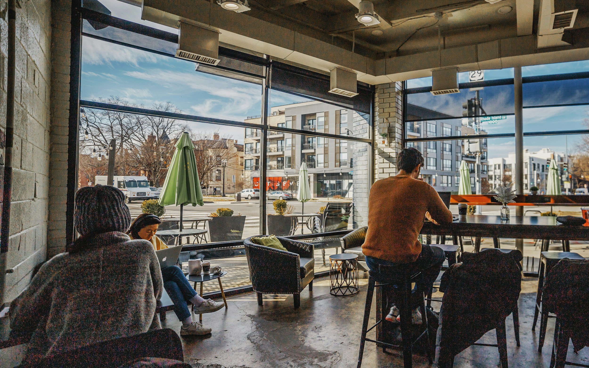 Interior of a modern coffee shop with large windows showing street view, customers sitting and working on laptops.