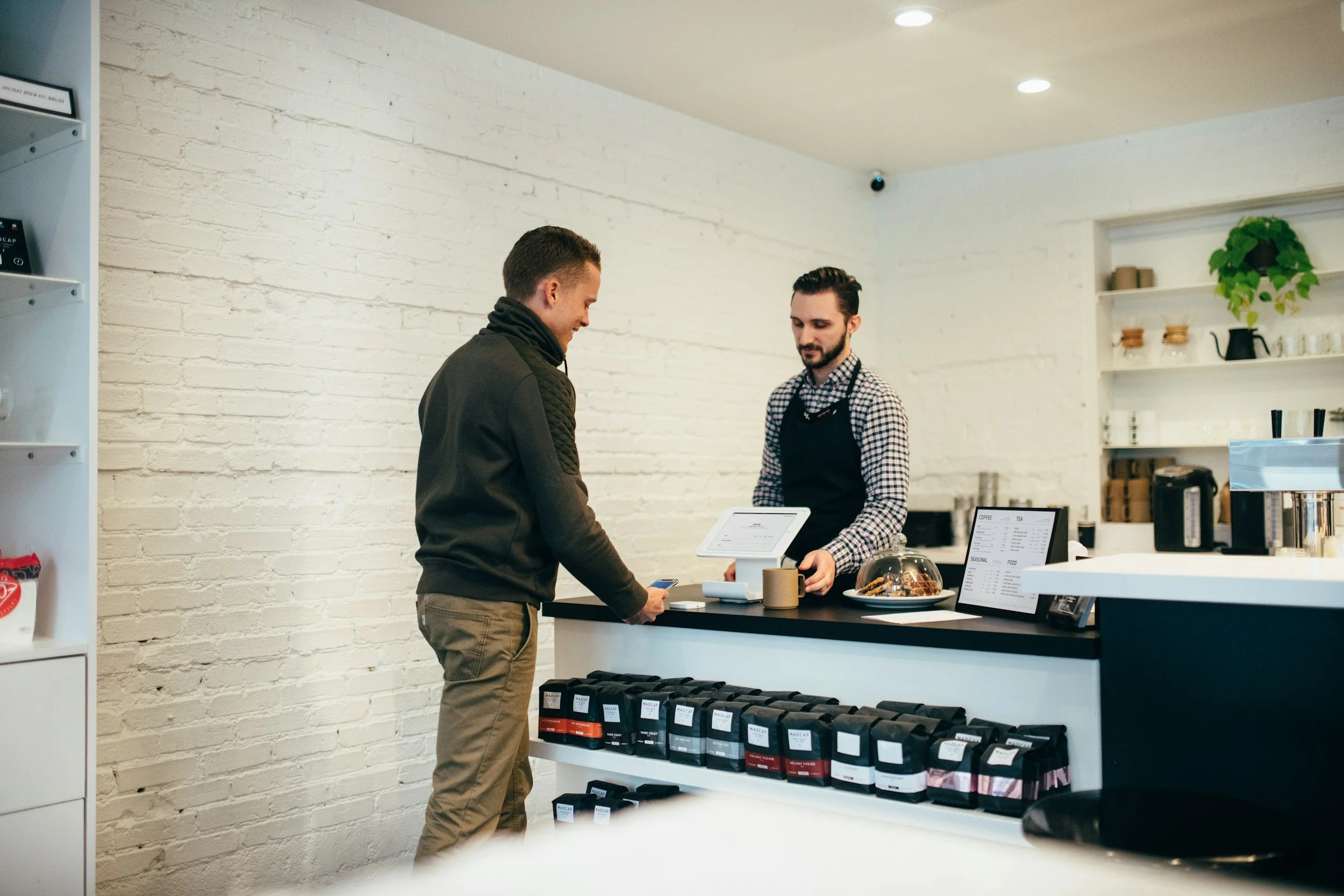 Customer paying at coffee shop counter with barista taking order, shelves with coffee equipment and products in background.