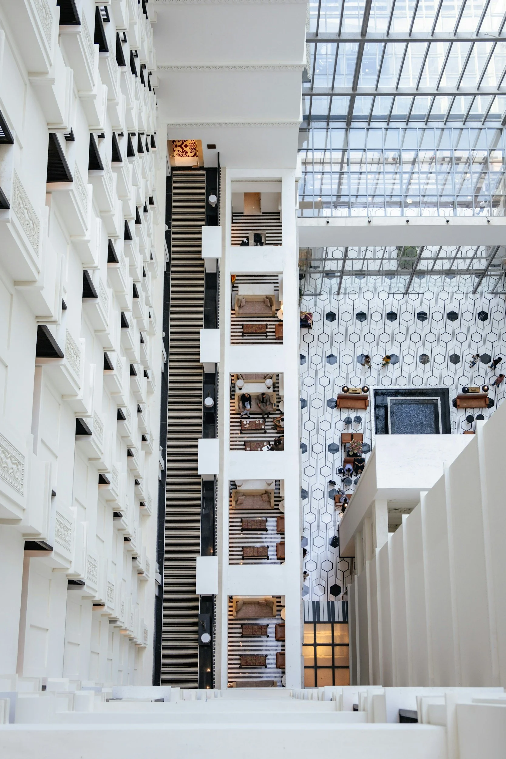 High-angle view of a modern hotel interior showing a central atrium, with multiple floors of white balconies, a patterned tiled floor, and a glass ceiling.