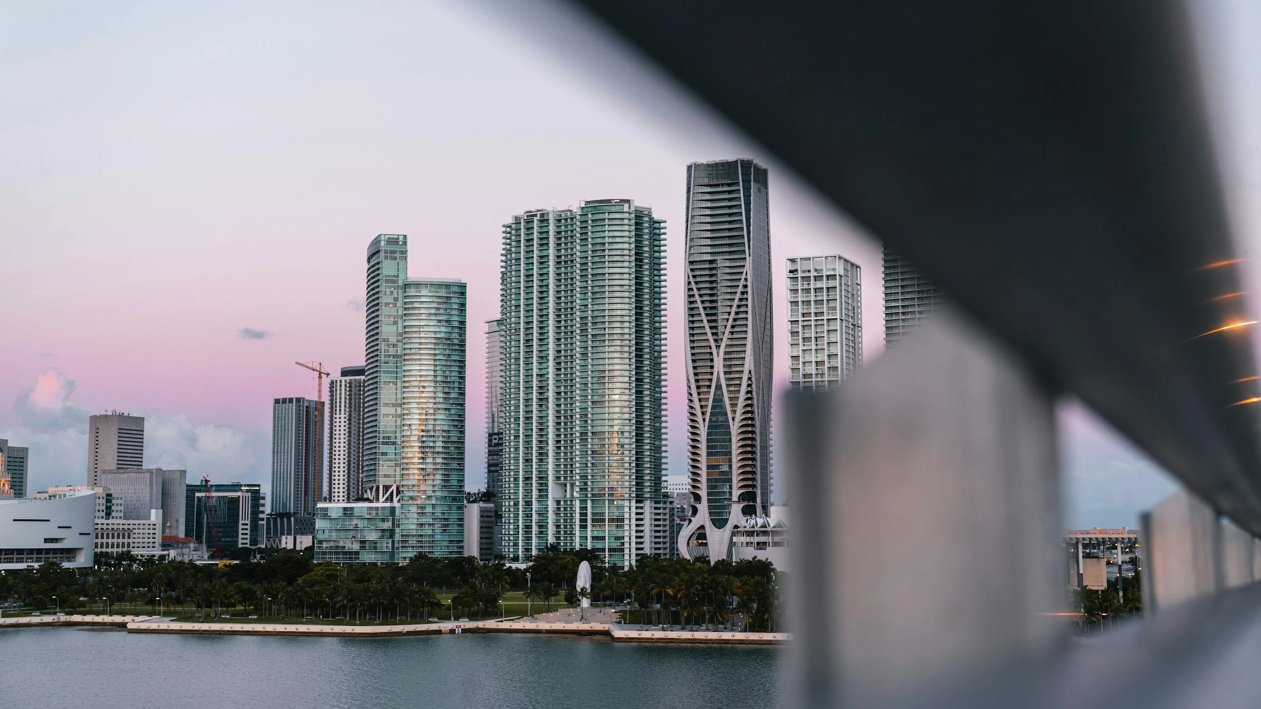 A city skyline with tall modern skyscrapers viewed from a waterfront park, partially obscured by a dark diagonal structural element in the foreground, during dusk with a pinkish sky.