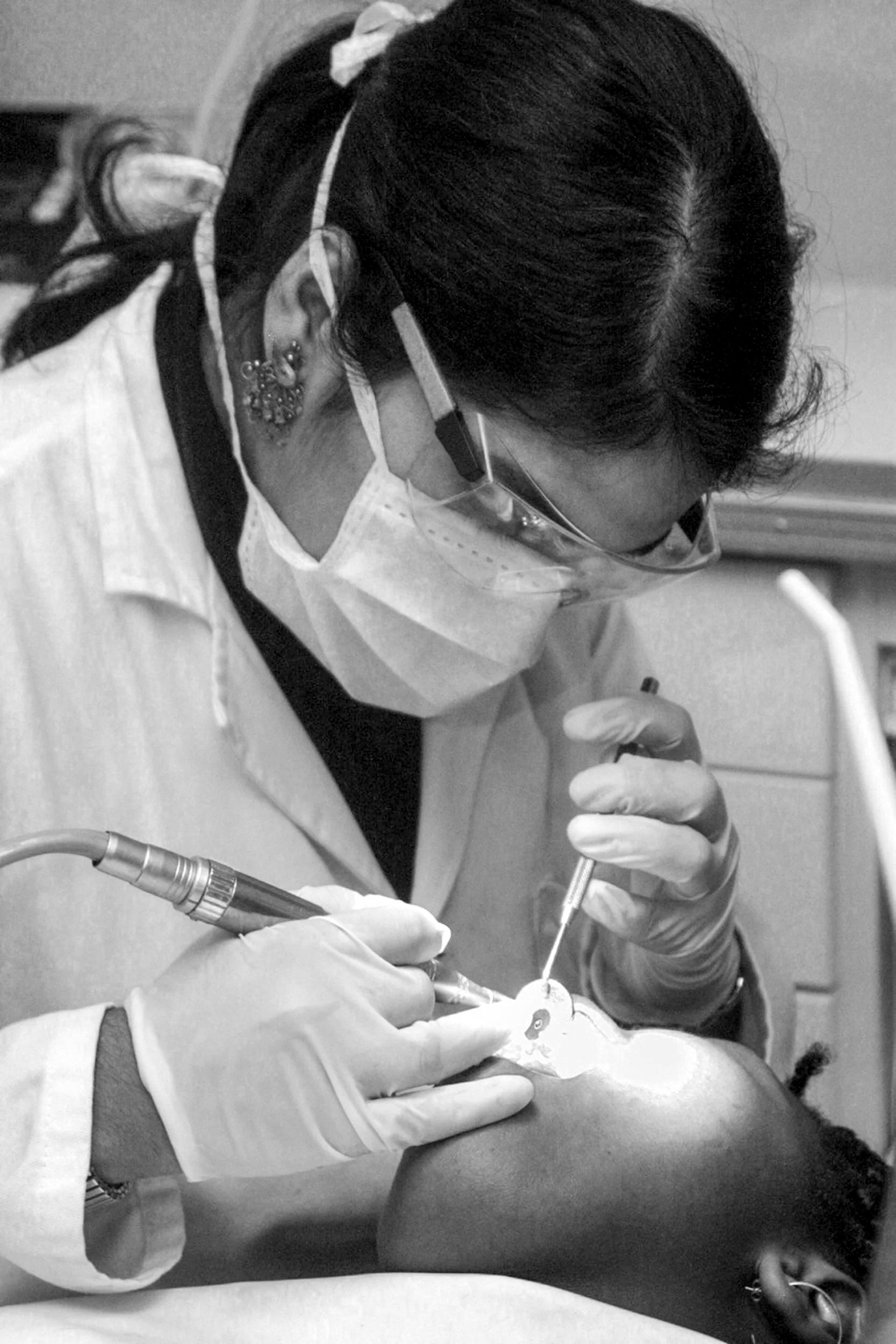 A female healthcare professional performing a dental procedure on a patient, wearing protective gear including glasses, a mask, and gloves.