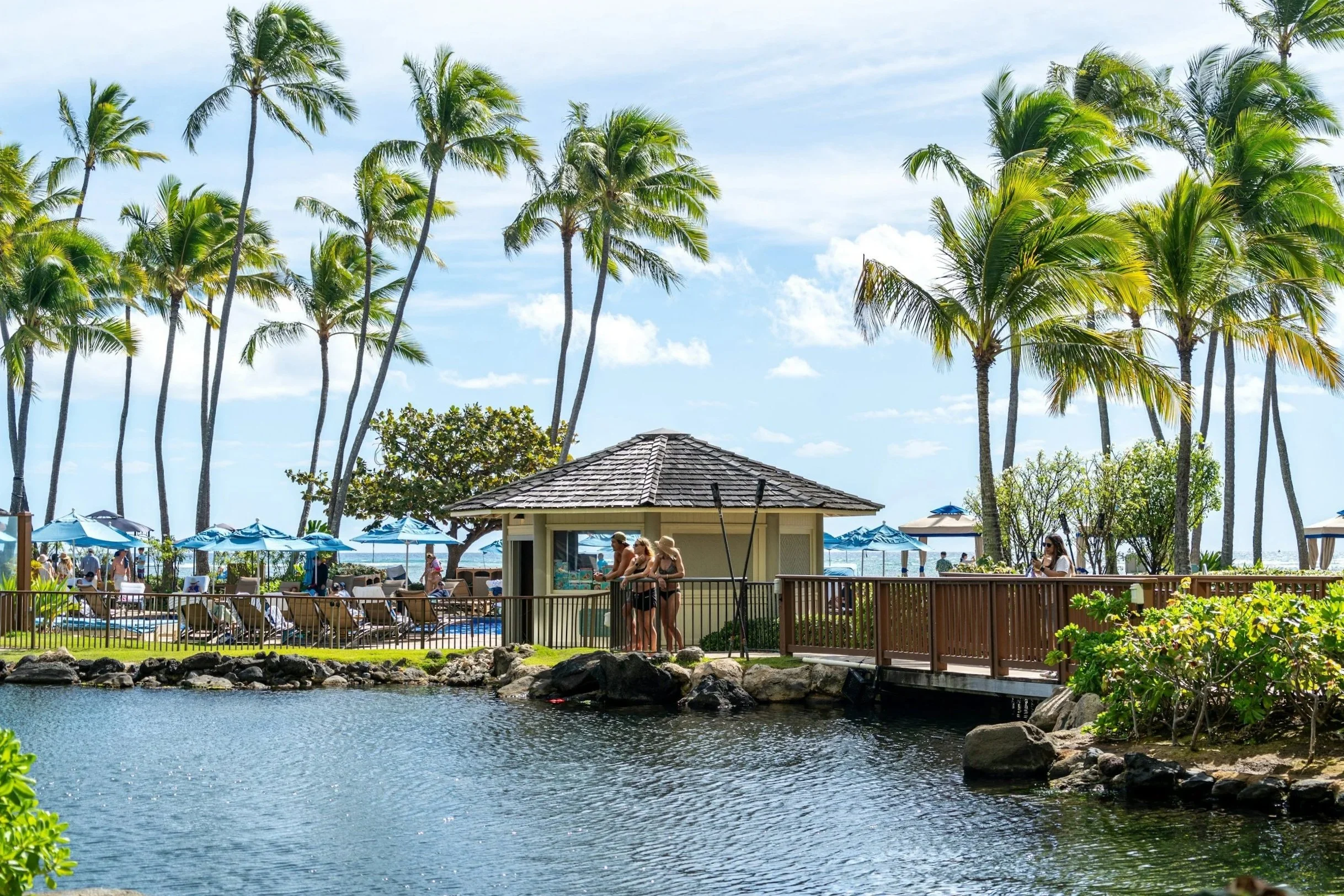 People by a small hut near a pond with palm trees and a beach with umbrellas in the background on a sunny day.