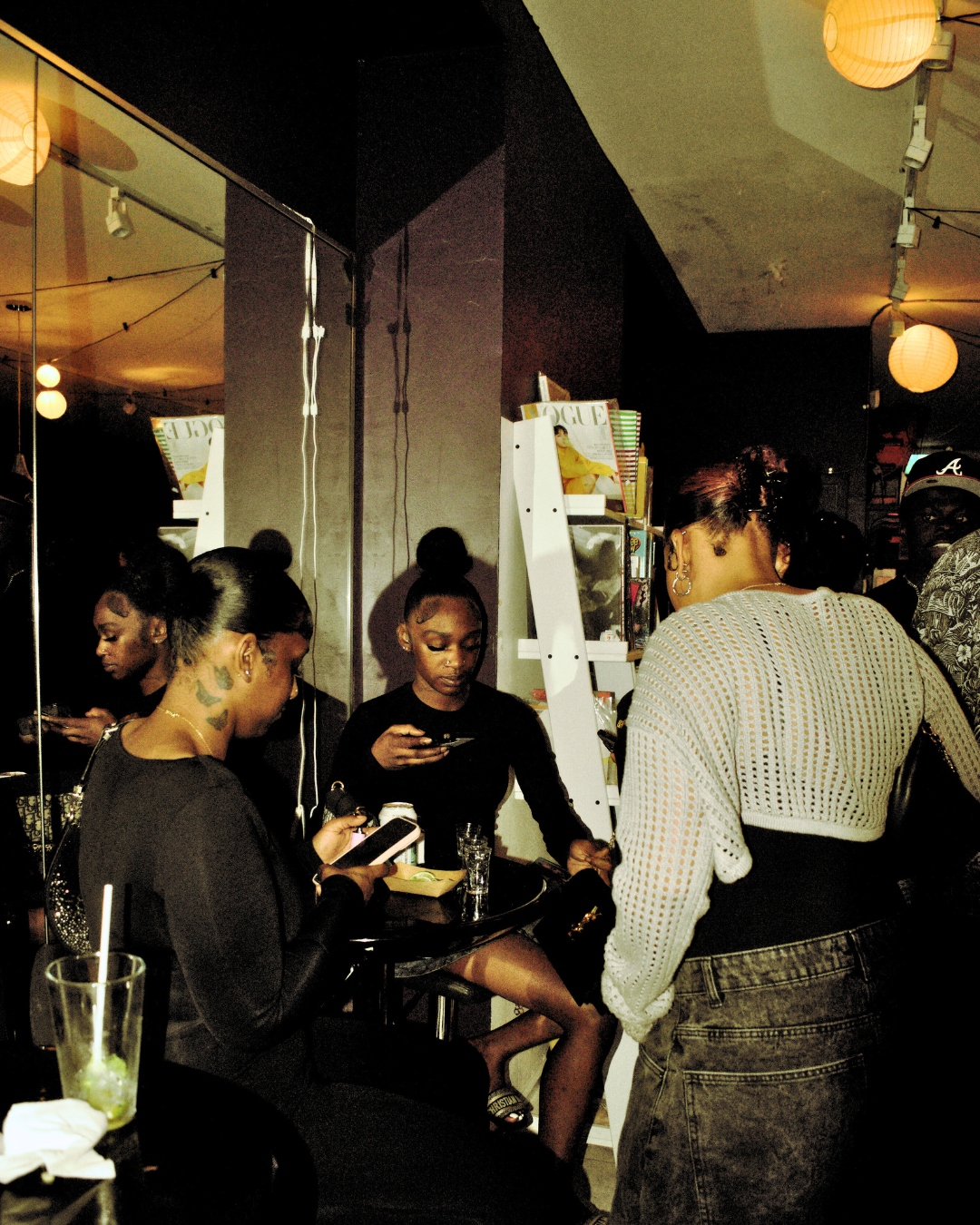 A group of young women socializing at a bar or cafe, using their phones, with books and magazines on shelves behind them and warm lighting.