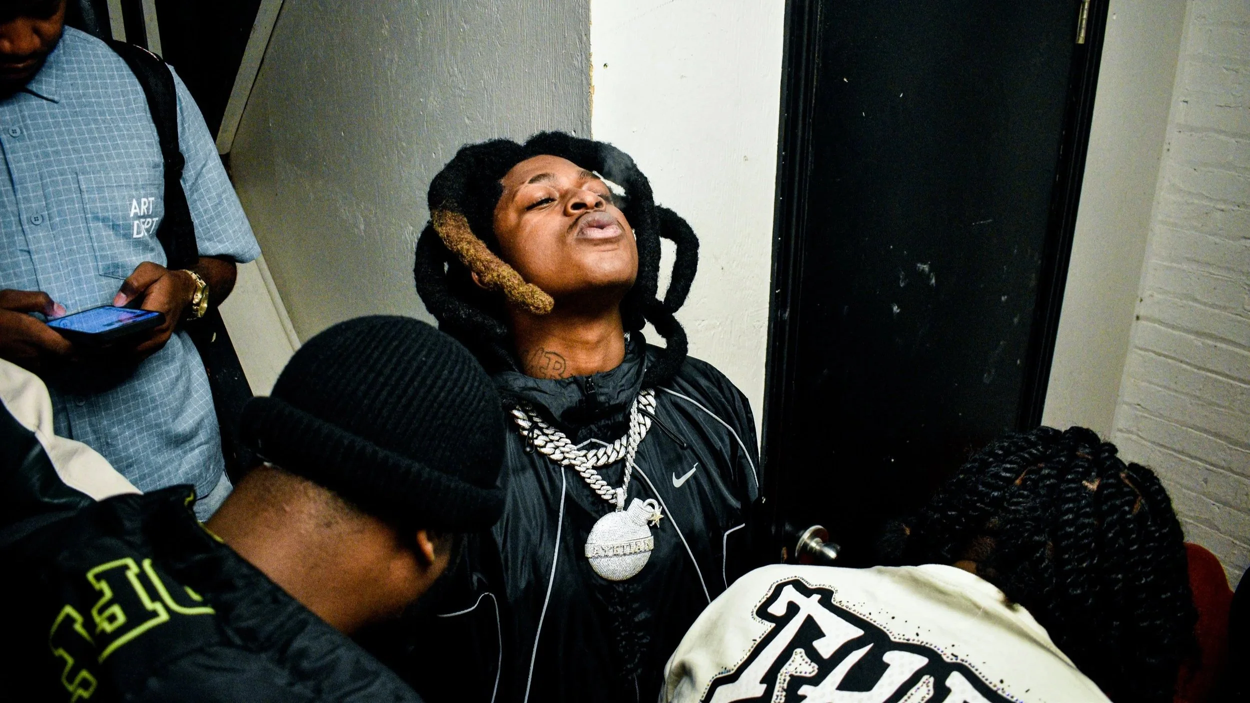 Ayetian, Dancehall artiste with styled dreadlocks and multiple chains around his neck, wearing a black jacket, appears to be in a moment of emotion with his head tilted back and eyes closed, seated with other people around him pre-show in Toronto.