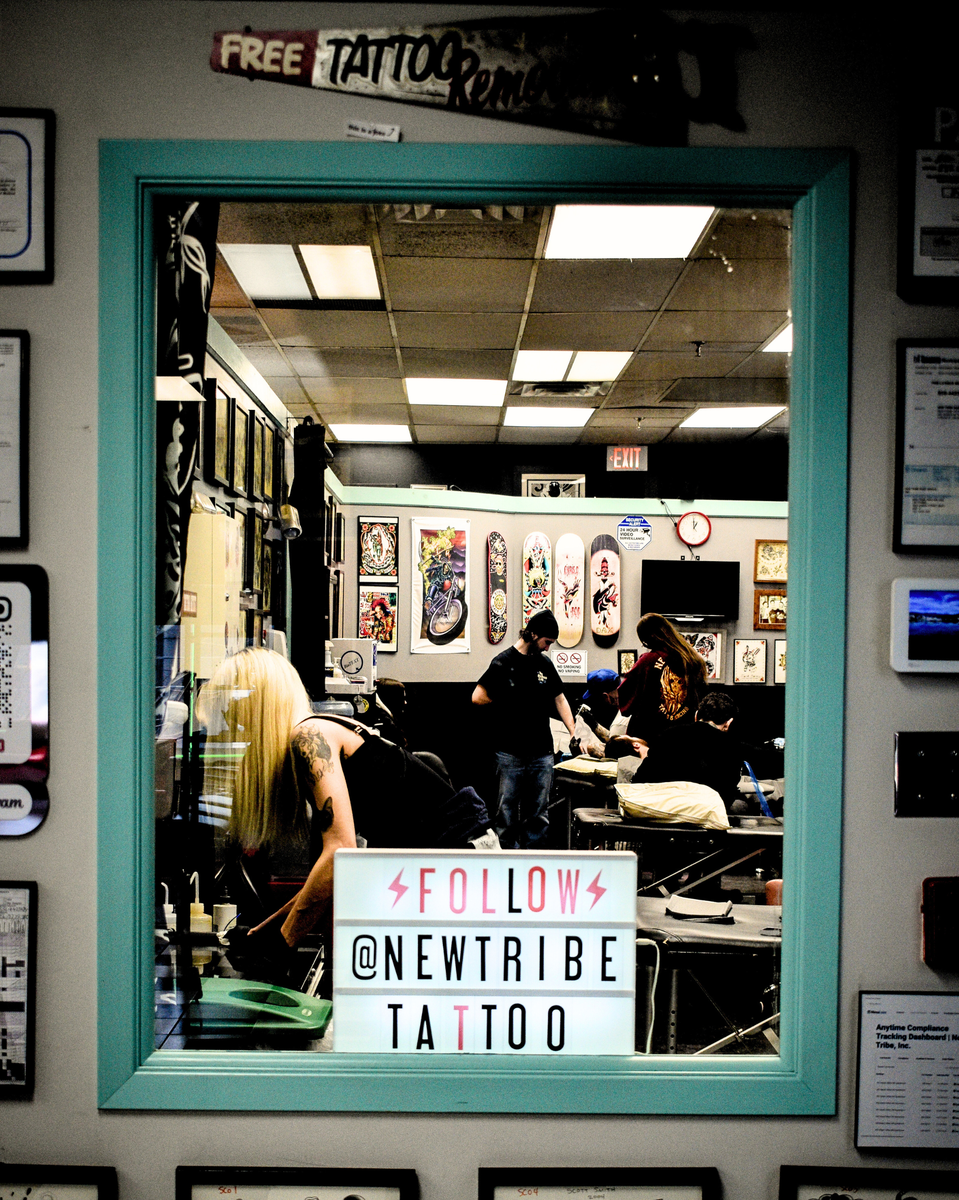 View through a window into a tattoo parlor with multiple people, tattoo artwork on the walls, and a sign that says 'Follow @NewTribeTattoo'.