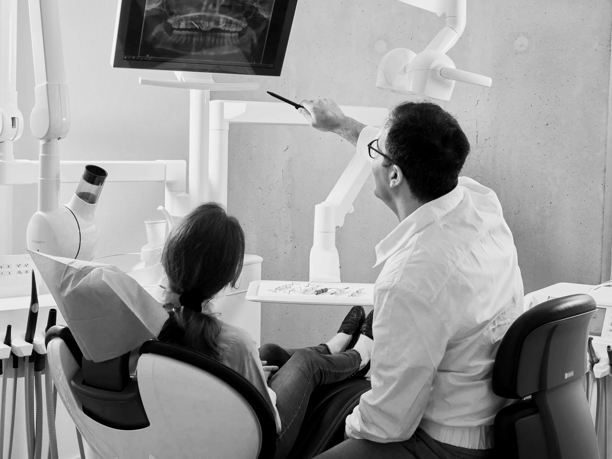 A dental consultation in a clinic where a dentist is pointing at an X-ray on a monitor while a young girl patient sits in the dental chair, wearing a dental bib.