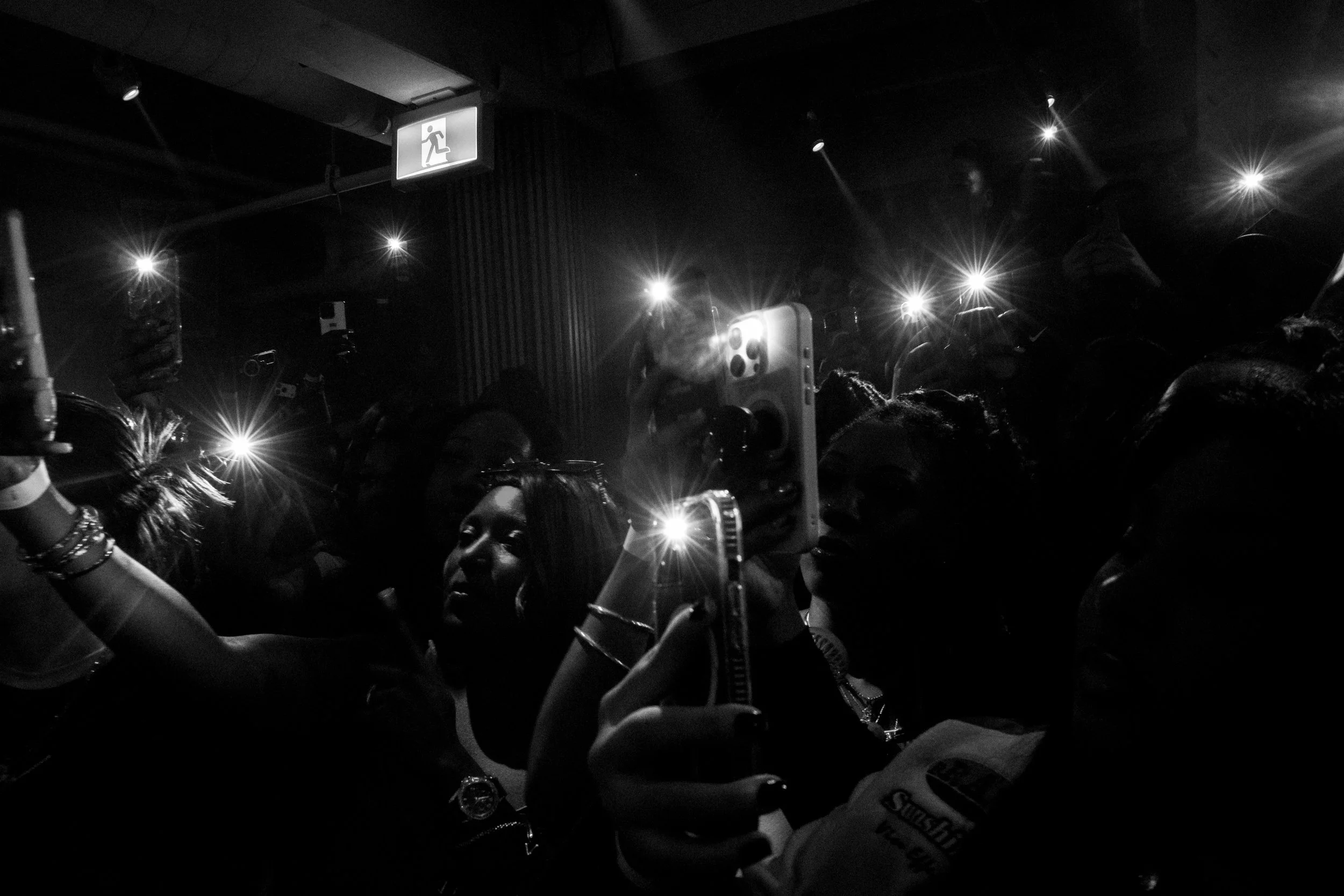 A group of people in a dark, crowded space taking photos with their phones at a show, with flashes illuminating the scene.