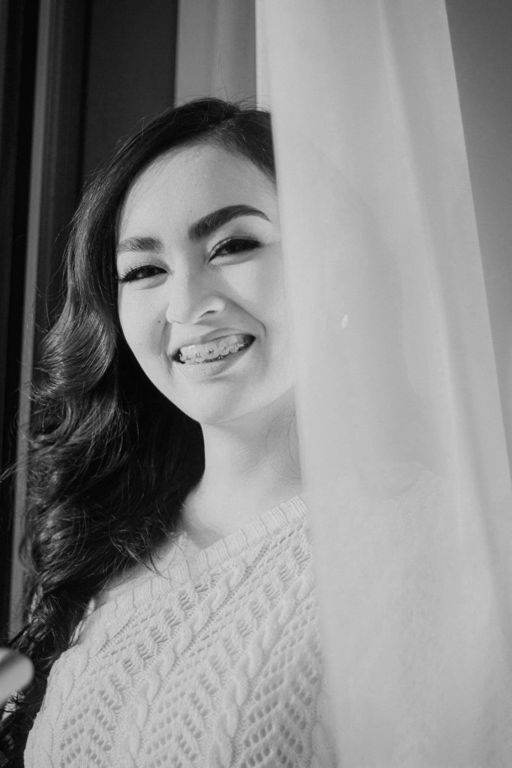 A young woman with dark, wavy hair, smiling and showing braces, peeking from behind a curtain.