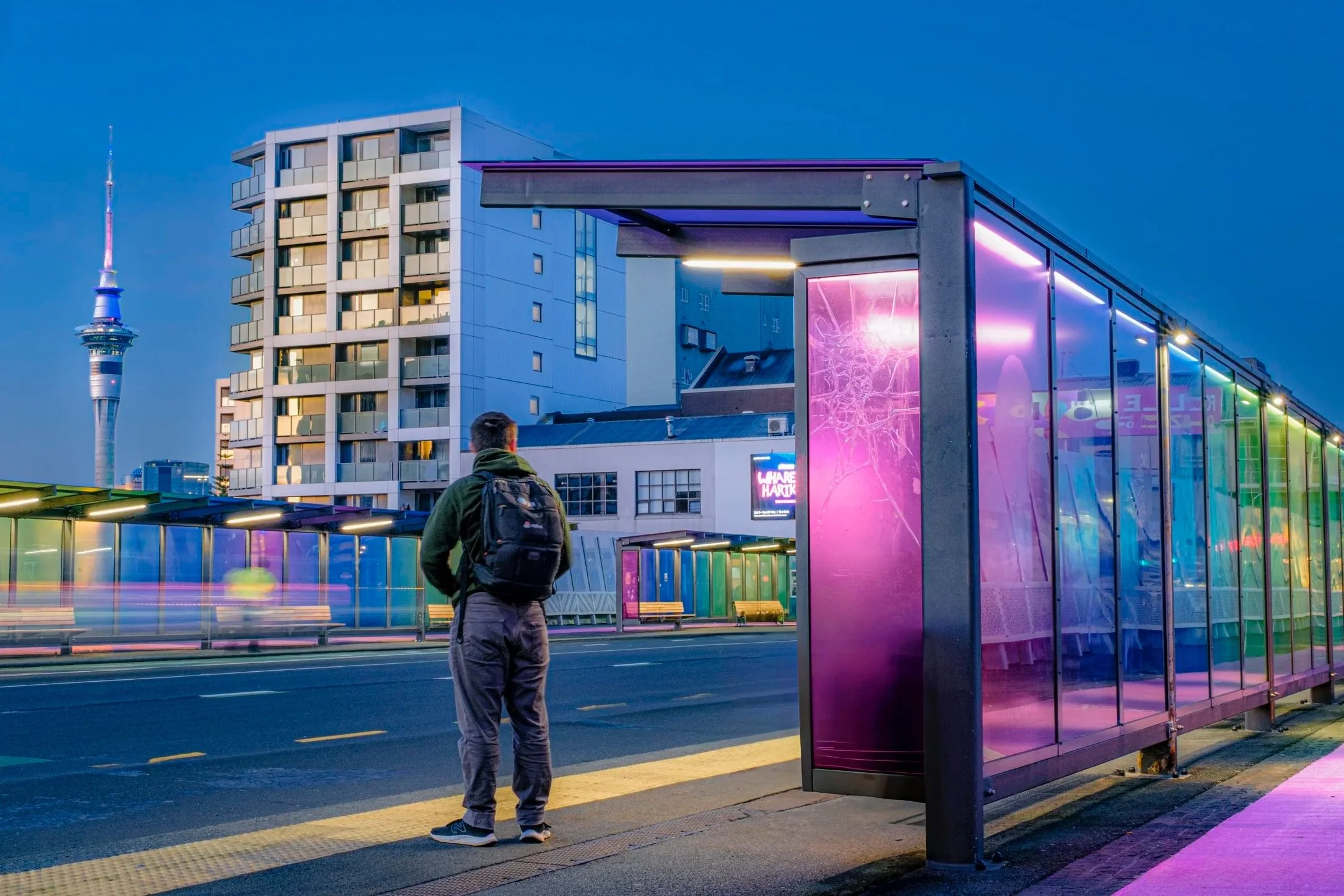 A person with a backpack standing at a bus stop with colourful neon lights, a modern building, and the Sky Tower, Auckland NZ in the background during early evening.
