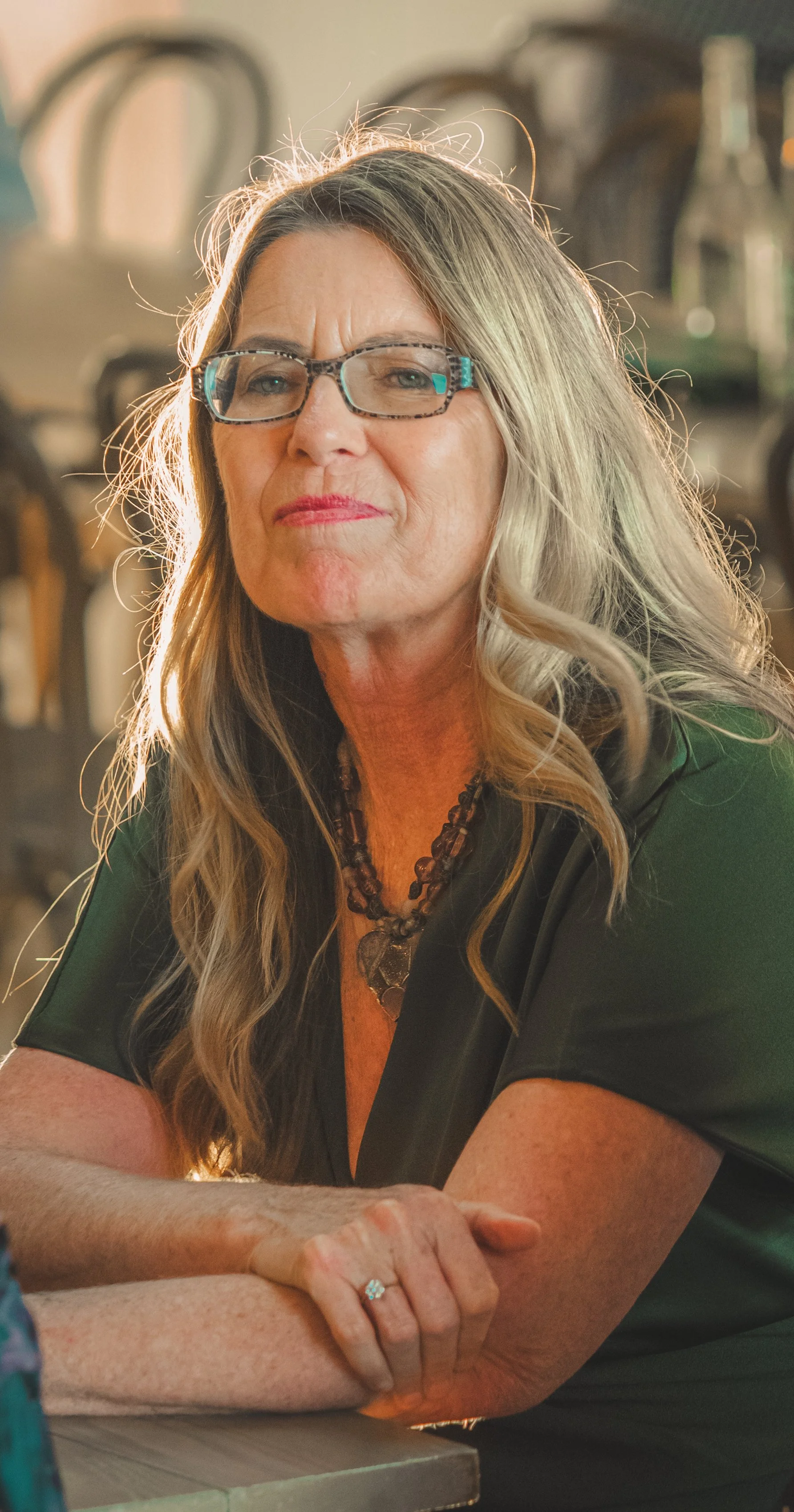A living portrait of a woman with long wavy blonde hair, wearing glasses, a black top, a beaded necklace, and a ring, sitting at a table with her arms crossed, looking at the camera. The background is blurred.