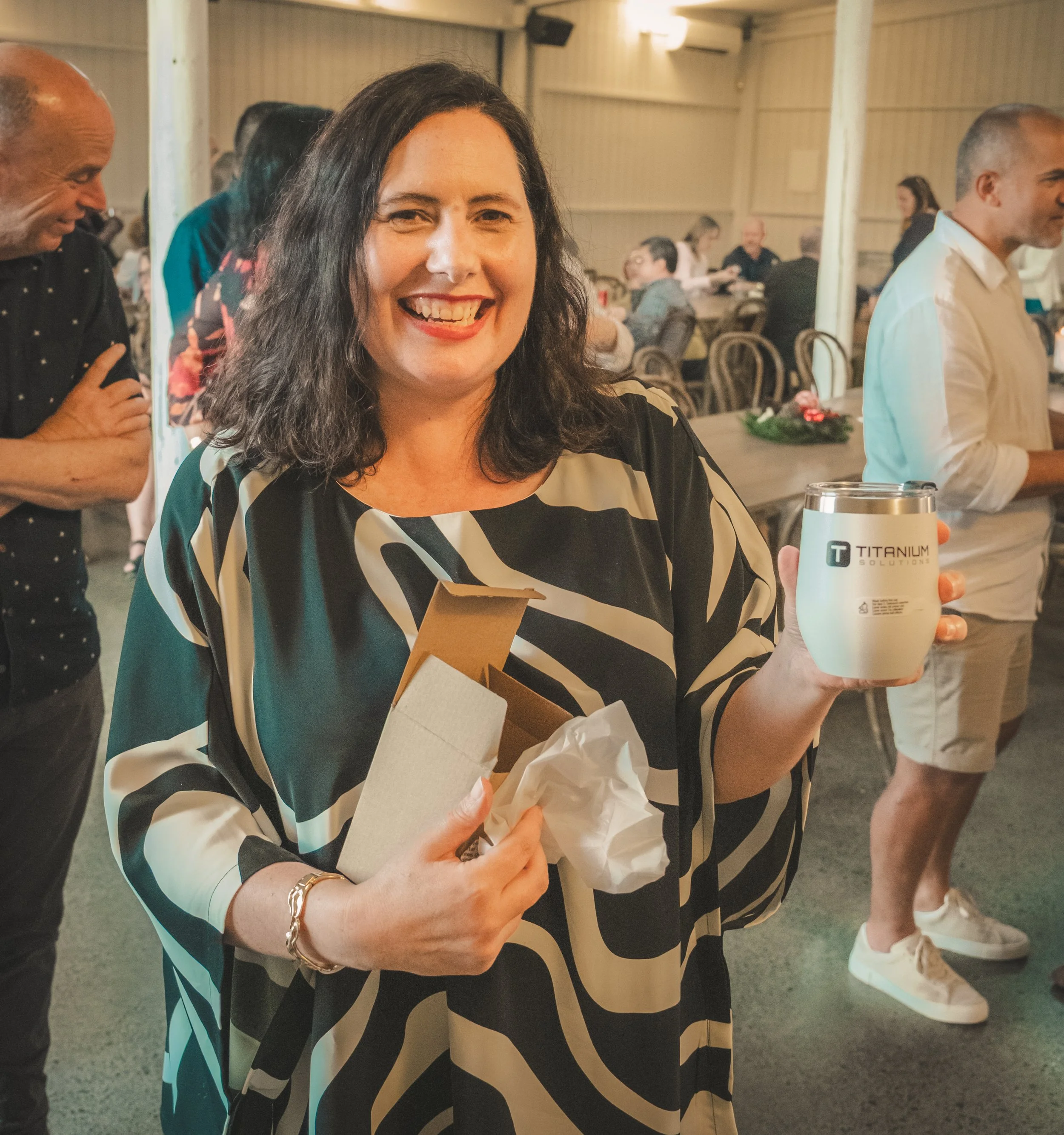 A woman with dark, curly hair and a broad smile holding a boxed gift and a tumbler in her hands at a social gathering indoors. She is wearing a black, white, and teal patterned blouse and a gold bracelet. Other people are visible in the background, e