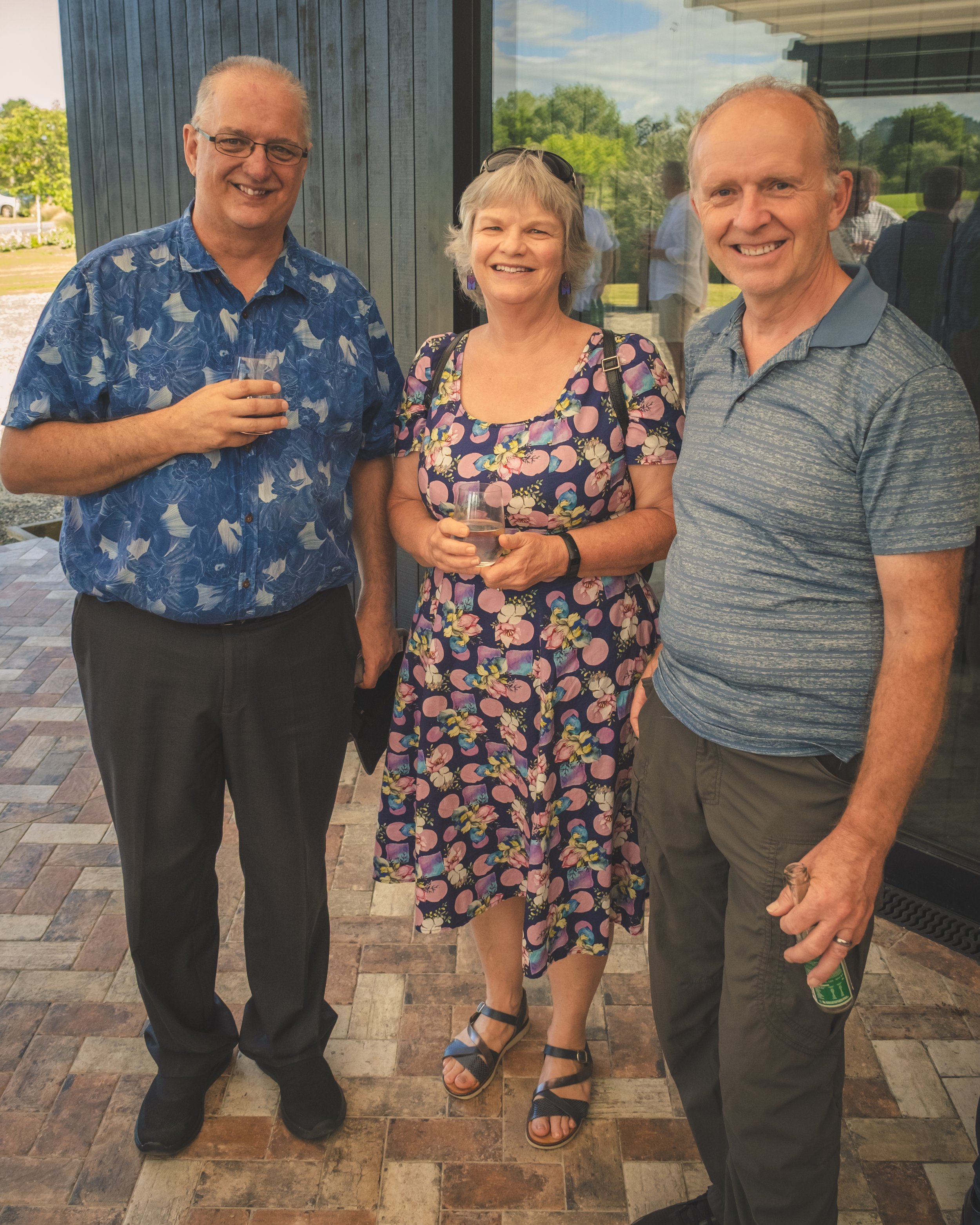 Three adults standing on a patio, smiling for a photo. The man on the left is wearing glasses and a blue patterned shirt, holding a glass of water. The woman in the middle is wearing a floral dress and sandals, also holding a glass of water. The man 