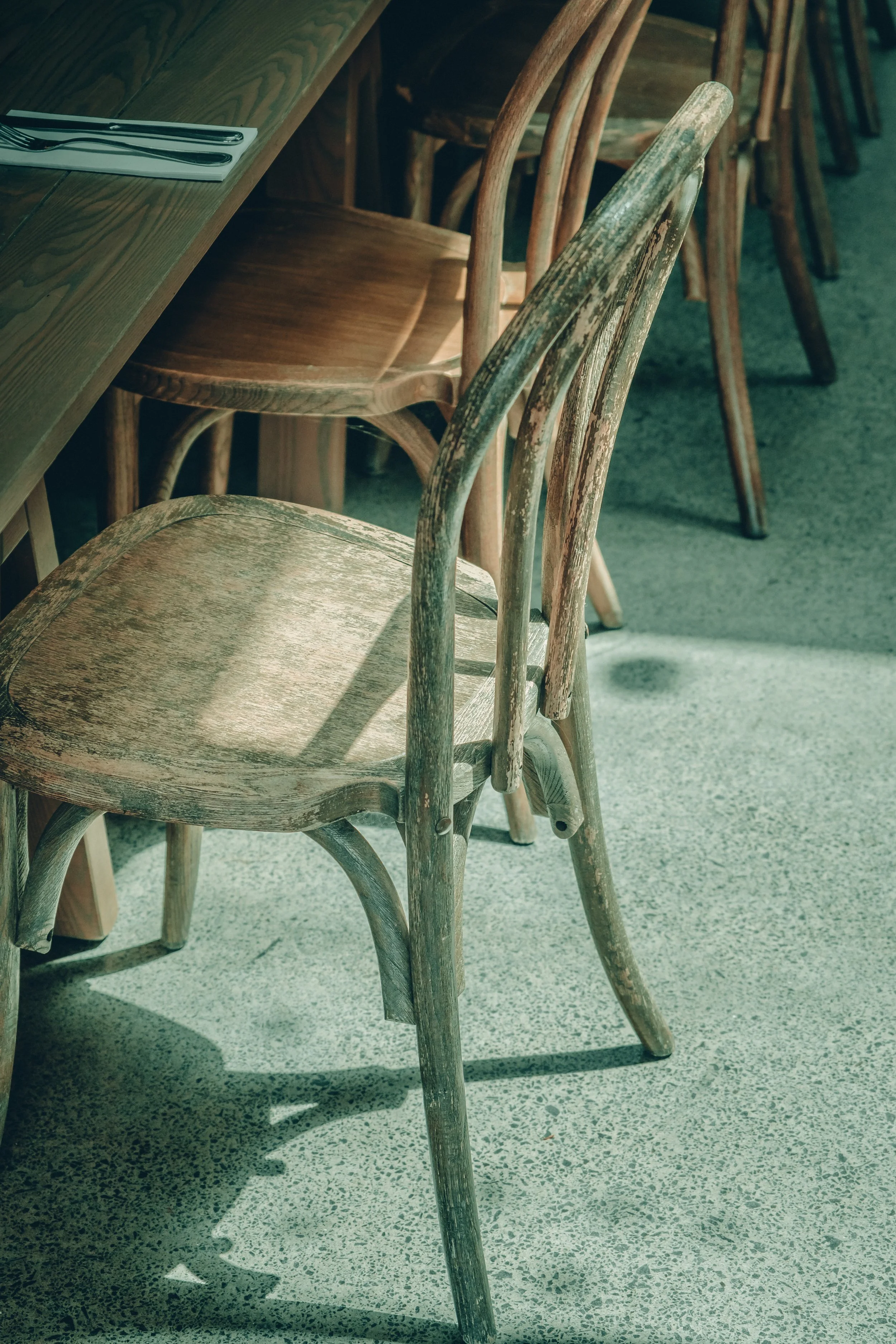 A row of weathered wooden chairs with rounded backs and slatted seats arranged along a wooden table in a restaurant or cafe setting, with shadows cast on the speckled gray floor.