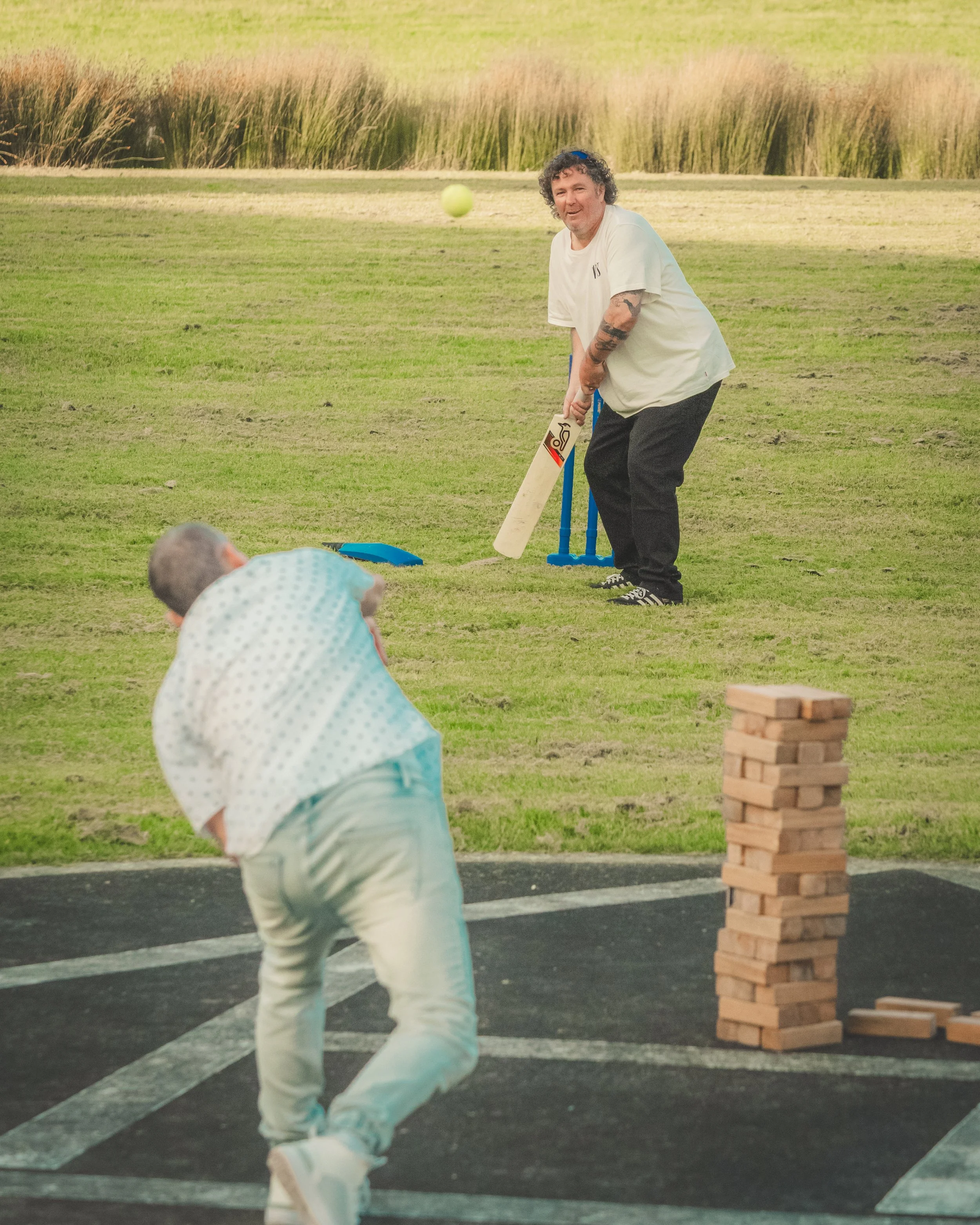 A man with curly hair and tattoos on his arms playing cricket outdoors, preparing to hit a yellow cricket ball. Children are watching in the foreground, and a stack of wooden blocks is nearby. The setting is a grassy field with a pathway and some tal