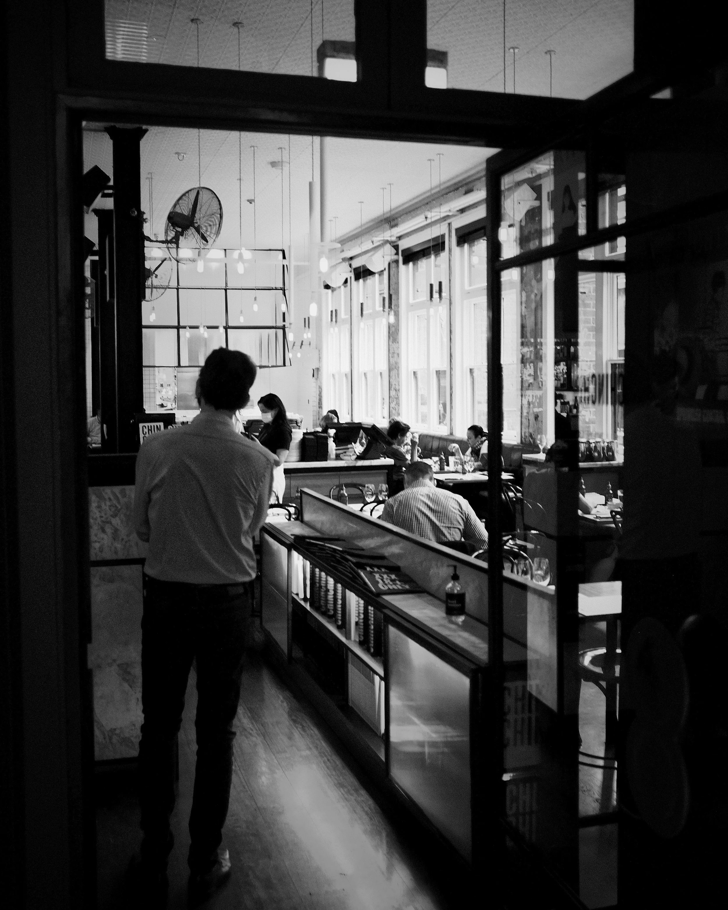 Black and white photo of a person walking into a cafe or restaurant, with large window panes allowing sunlight to fill the seating area. Several people are seated inside, some using laptops or reading. The interior features hanging lights and decorat