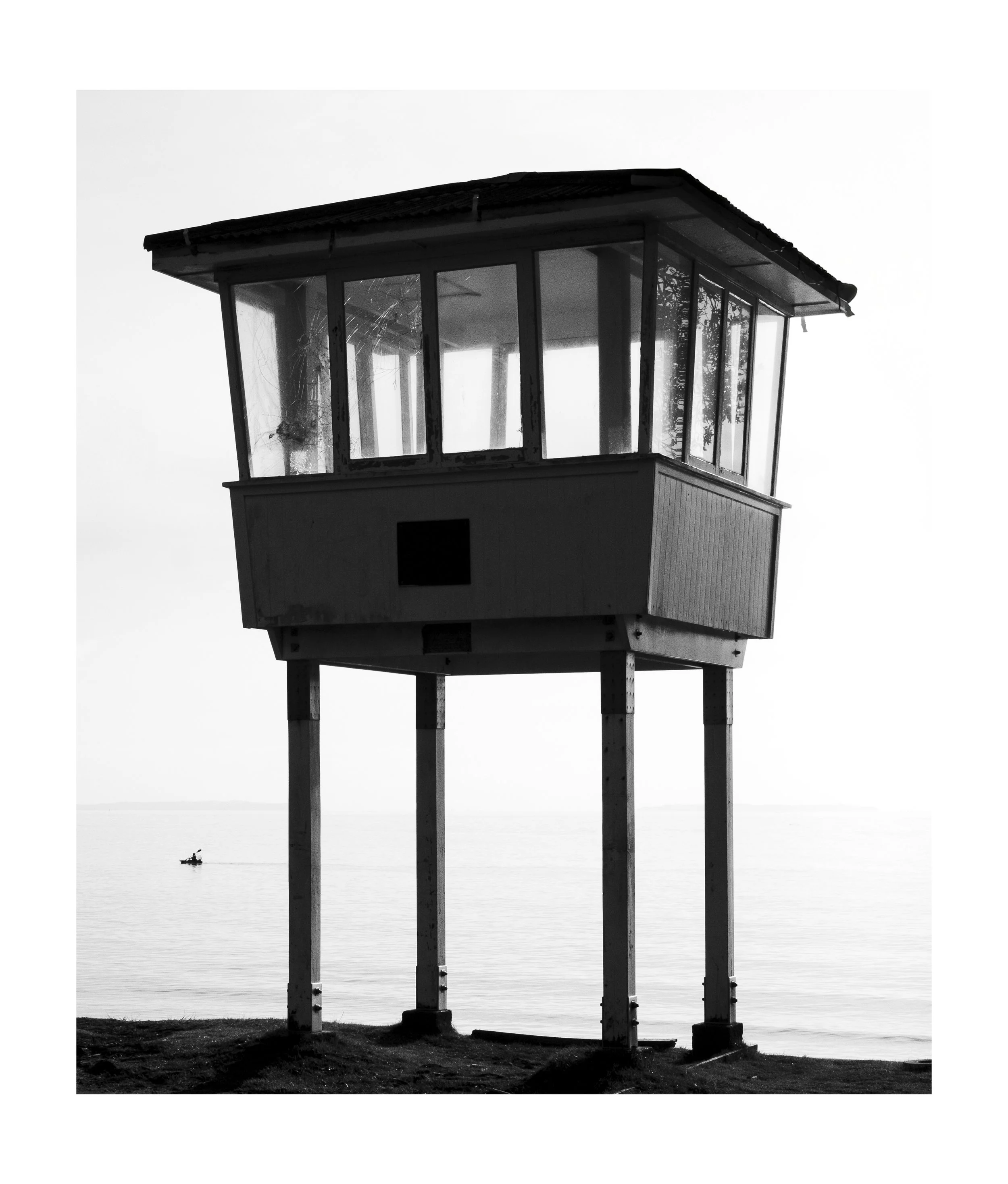 Black and white photo of a watchtower on stilts over the water, with a boat in the distance, and broken glass on the tower's window.