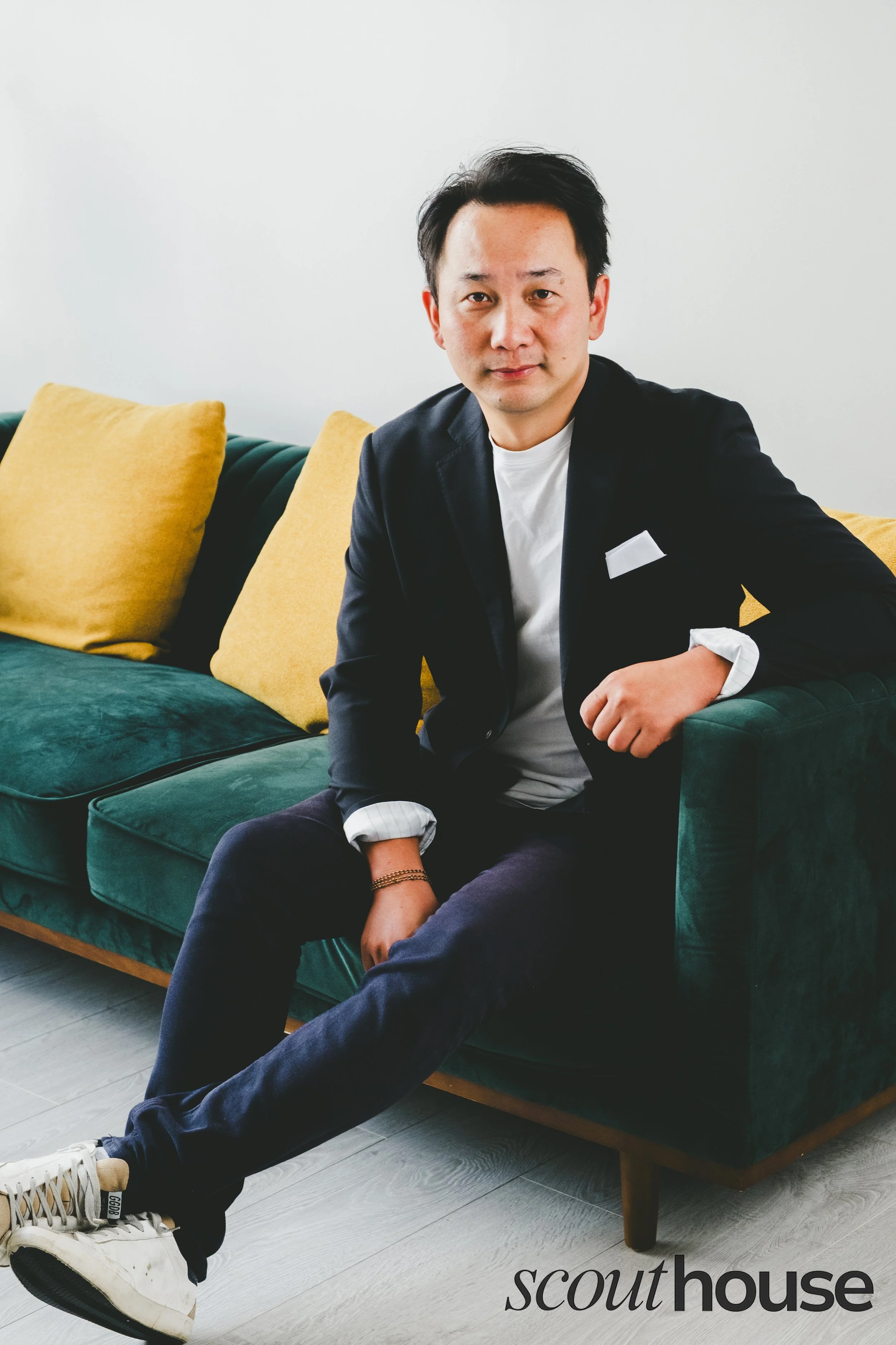 A man sitting on a green velvet couch with yellow pillows against a white wall. He is wearing a black blazer, white shirt, dark pants, and white sneakers, and is looking at the camera with a slight smile.