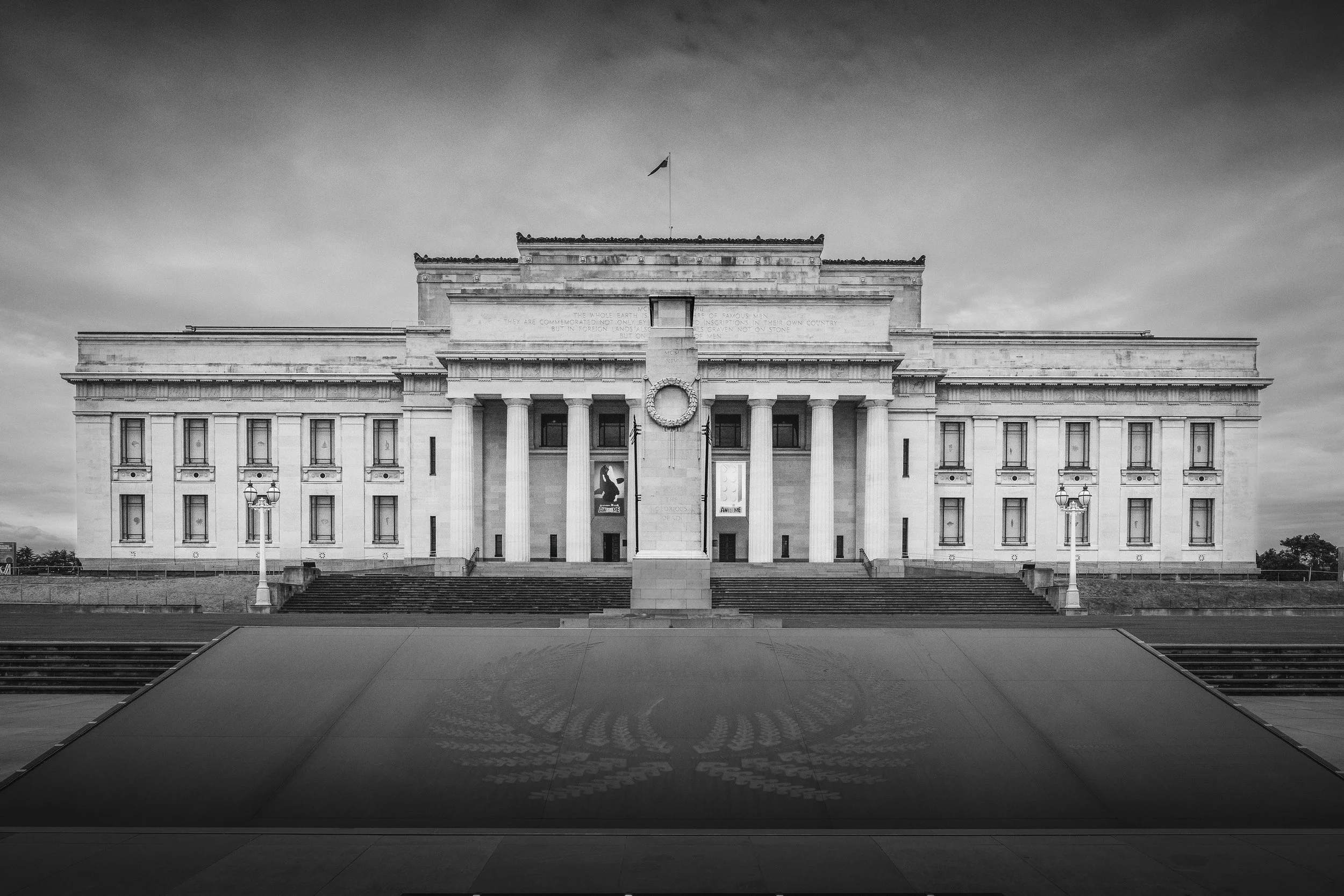 Black and white photo of the Auckland Museum, viewed from the front with stairs leading up to the monument and a cloudy sky in the background.