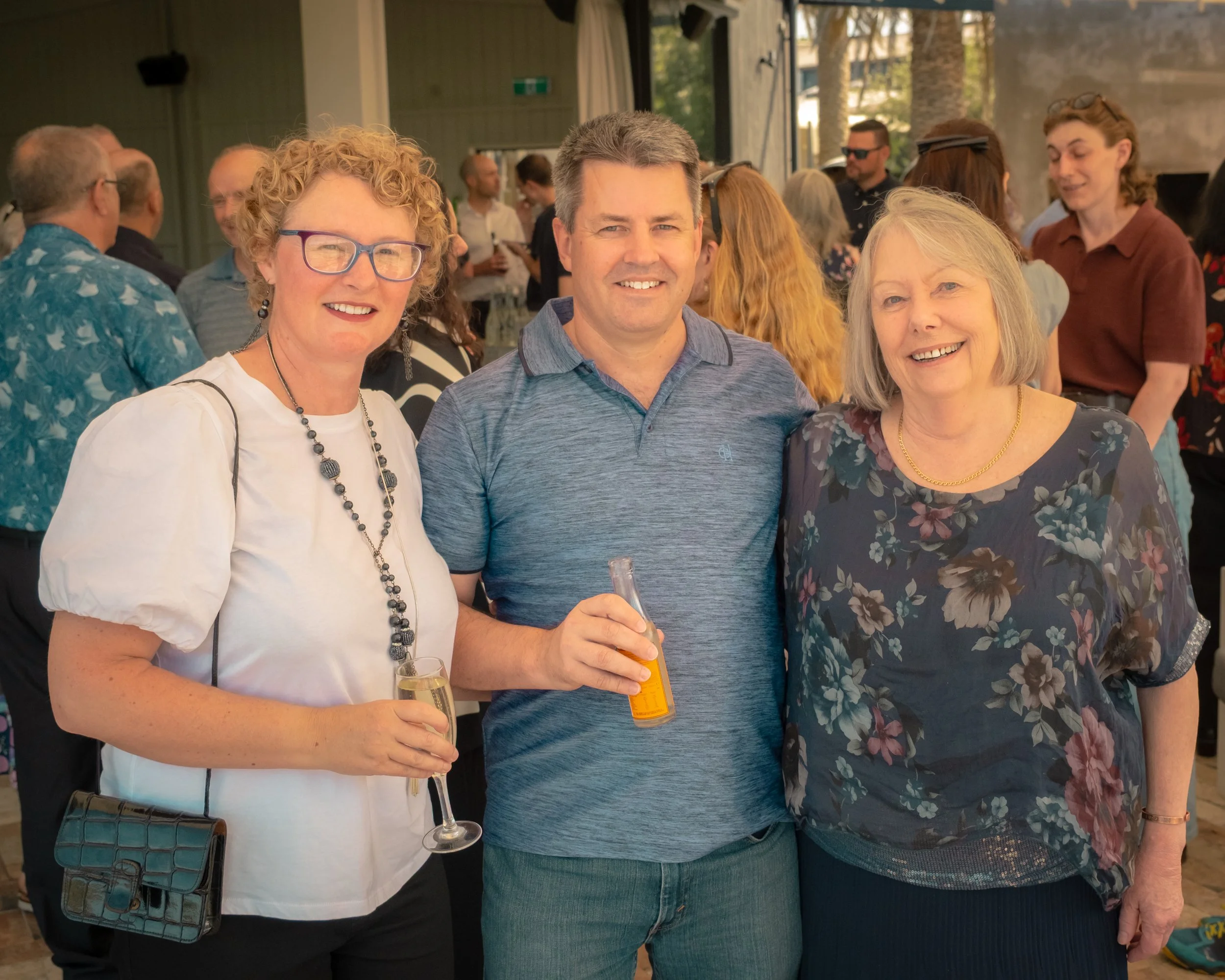 A group of people at a social gathering, with three women in the foreground smiling and holding drinks, surrounded by others in the background.