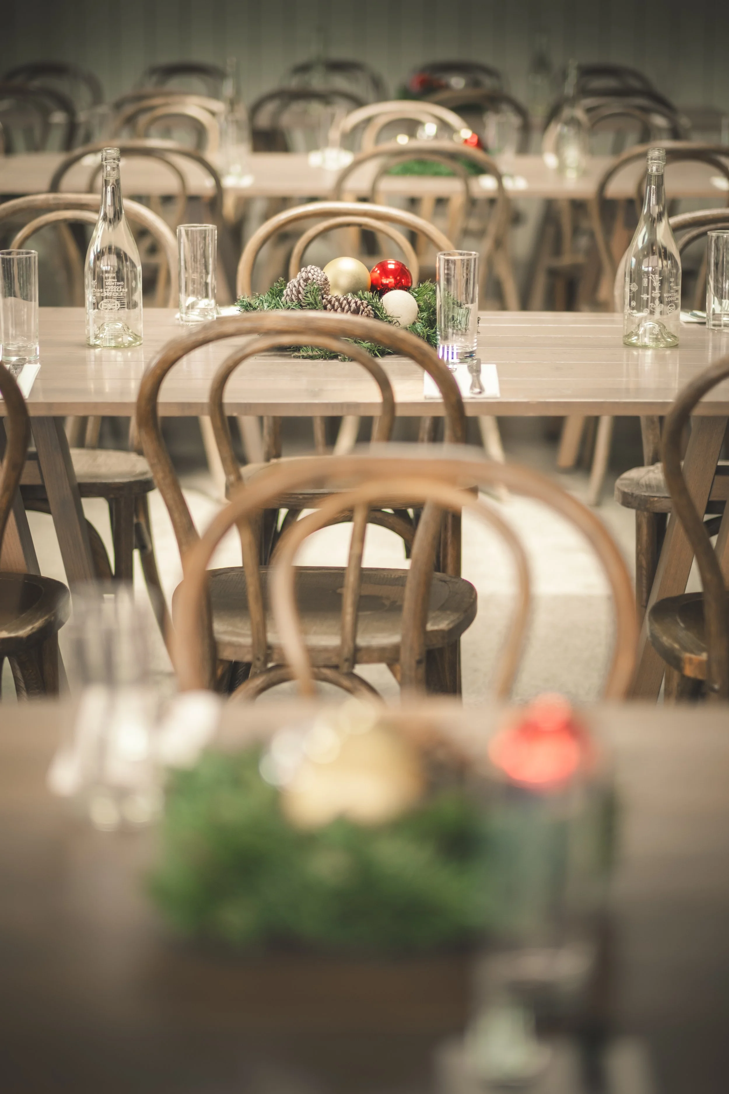 A long wooden dining table set for a holiday gathering with a centerpiece of Christmas ornaments and greenery, surrounded by wooden chairs, in a dimly lit room.