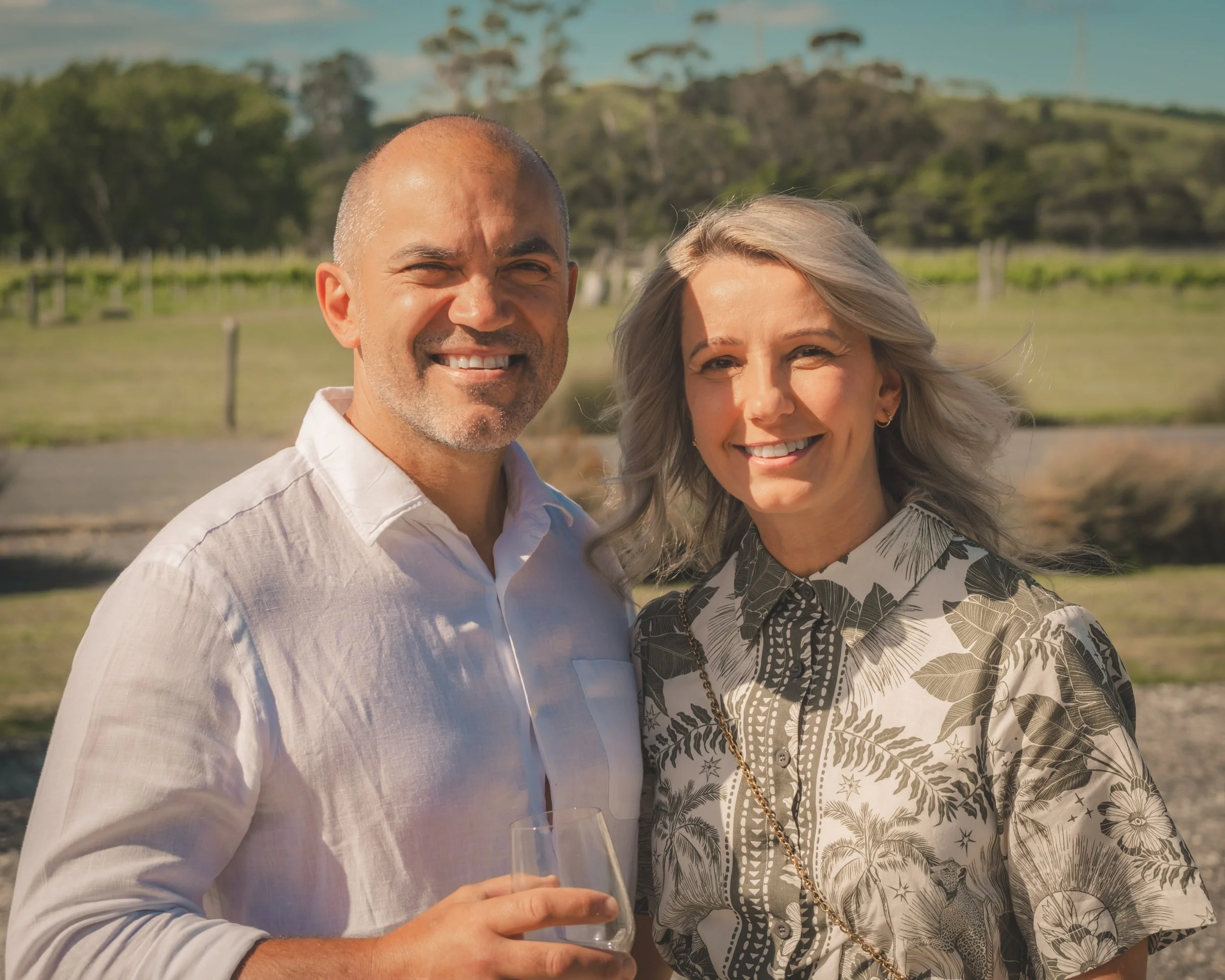 A smiling man in a white shirt and a woman in a patterned top standing outdoors on a sunny day. The man is holding a glass of wine, and they are standing close together with a green landscape and blue sky in the background.