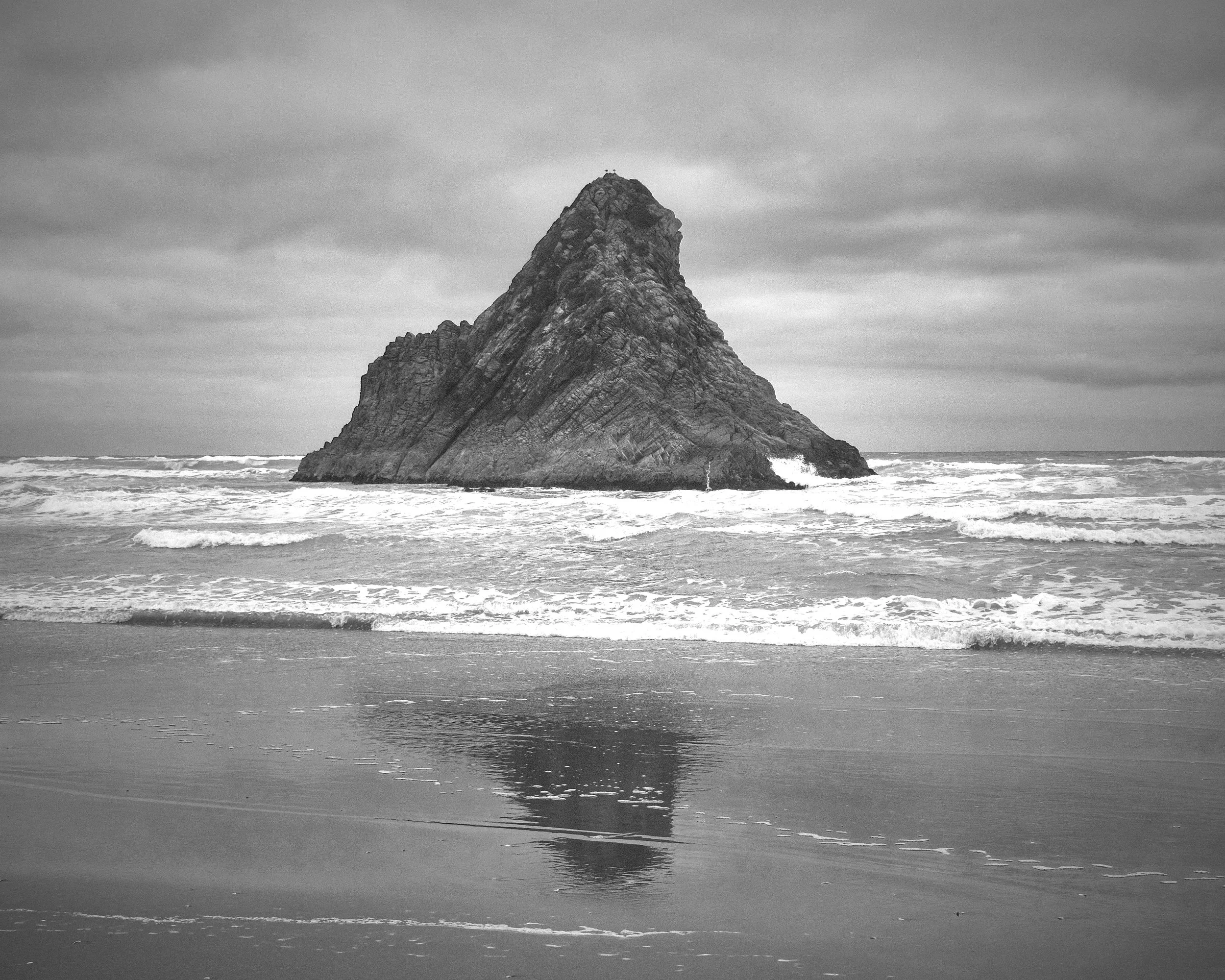 A black and white photograph of a large, jagged rock formation rising from the ocean with waves crashing against it and a cloudy sky overhead.