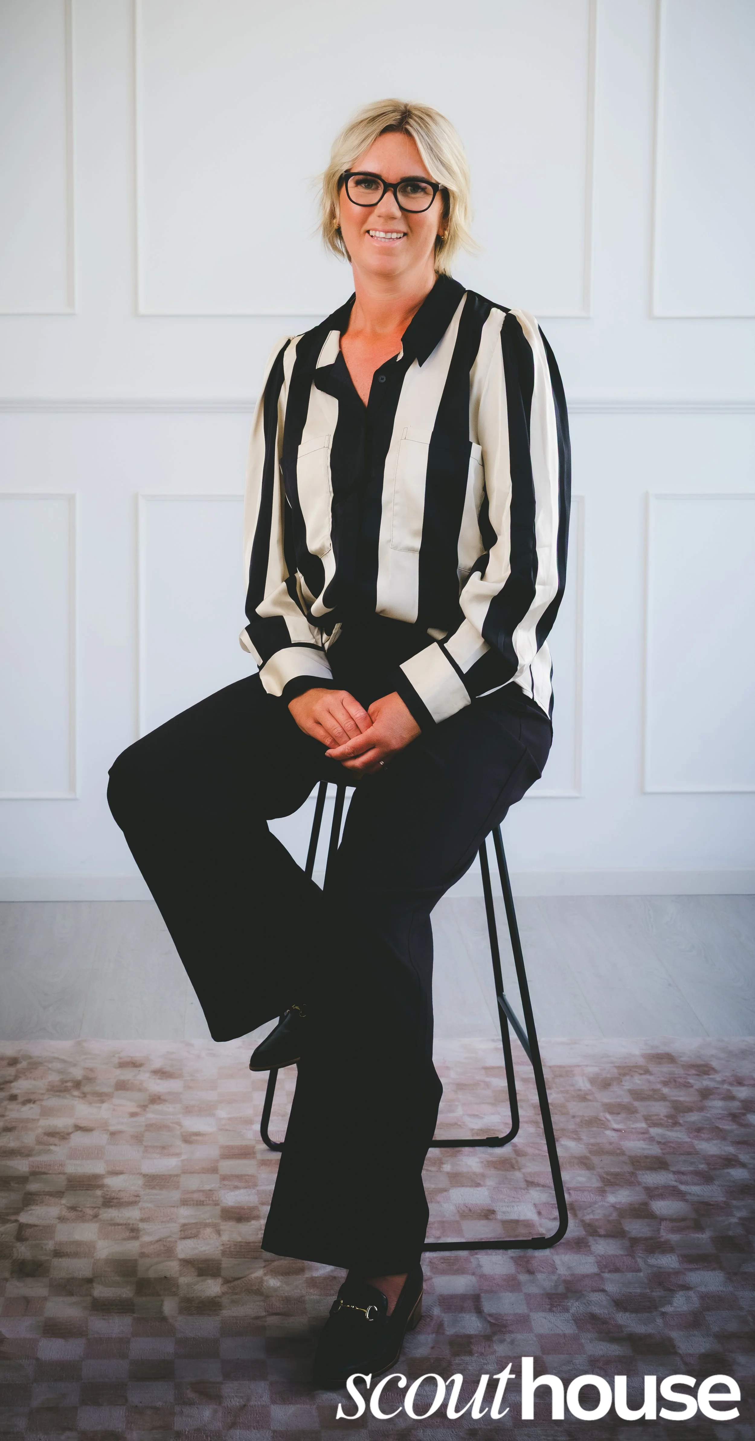 A woman with blonde hair, glasses, wearing a black and white striped blouse and black pants, sitting on a stool in a room with white panel walls and light-colored carpet, smiling at the camera.