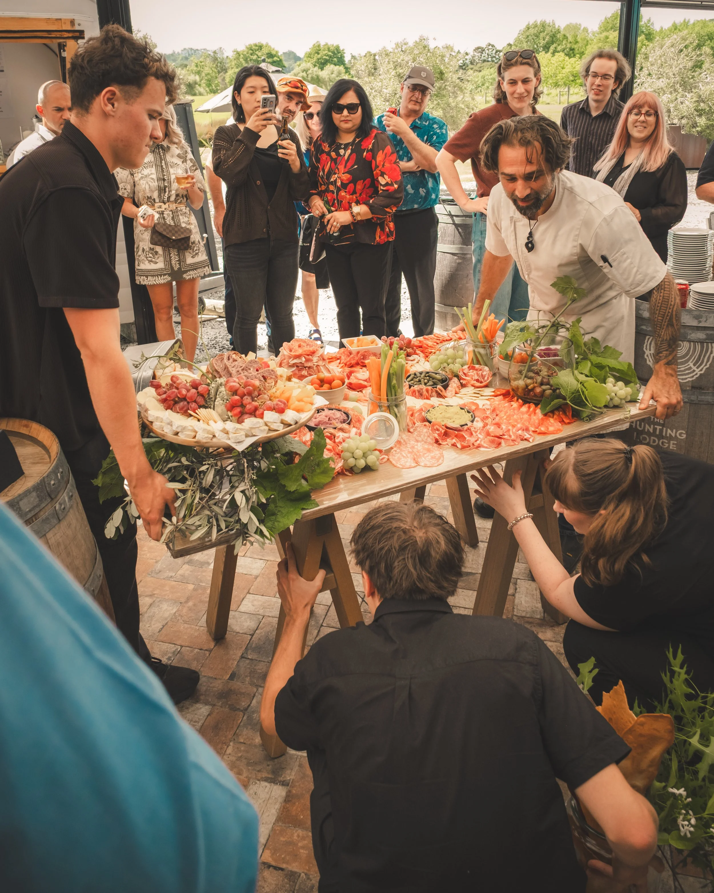 Group of people gathered around a table filled with various meats, cheeses, fruits, and vegetables during an outdoor event, with some taking photos and others observing.