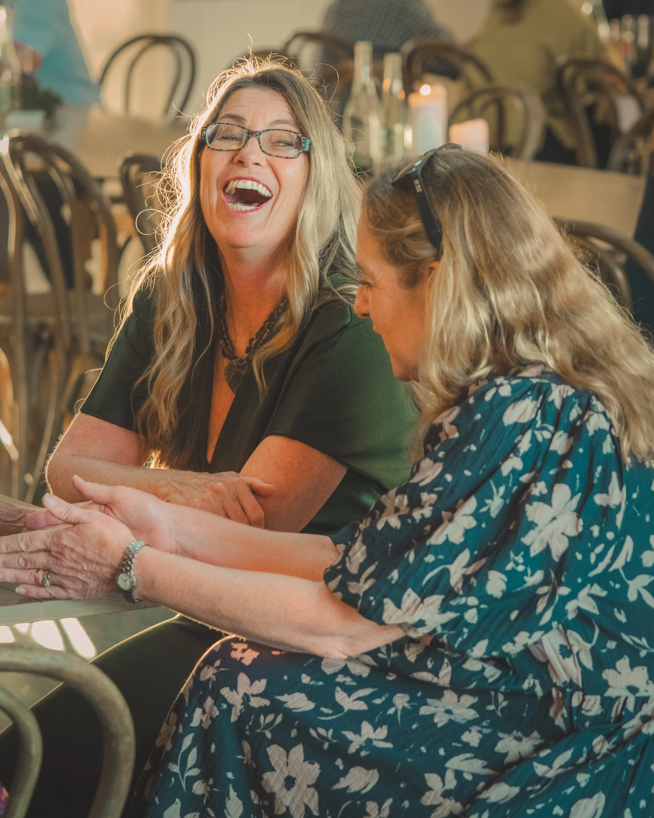 Two women sit together, laughing and holding hands. One woman has long blonde hair, wears glasses, and a green top. The other woman has short blonde hair, wears glasses, and a blue dress with a white floral pattern. They are in a restaurant with a bl