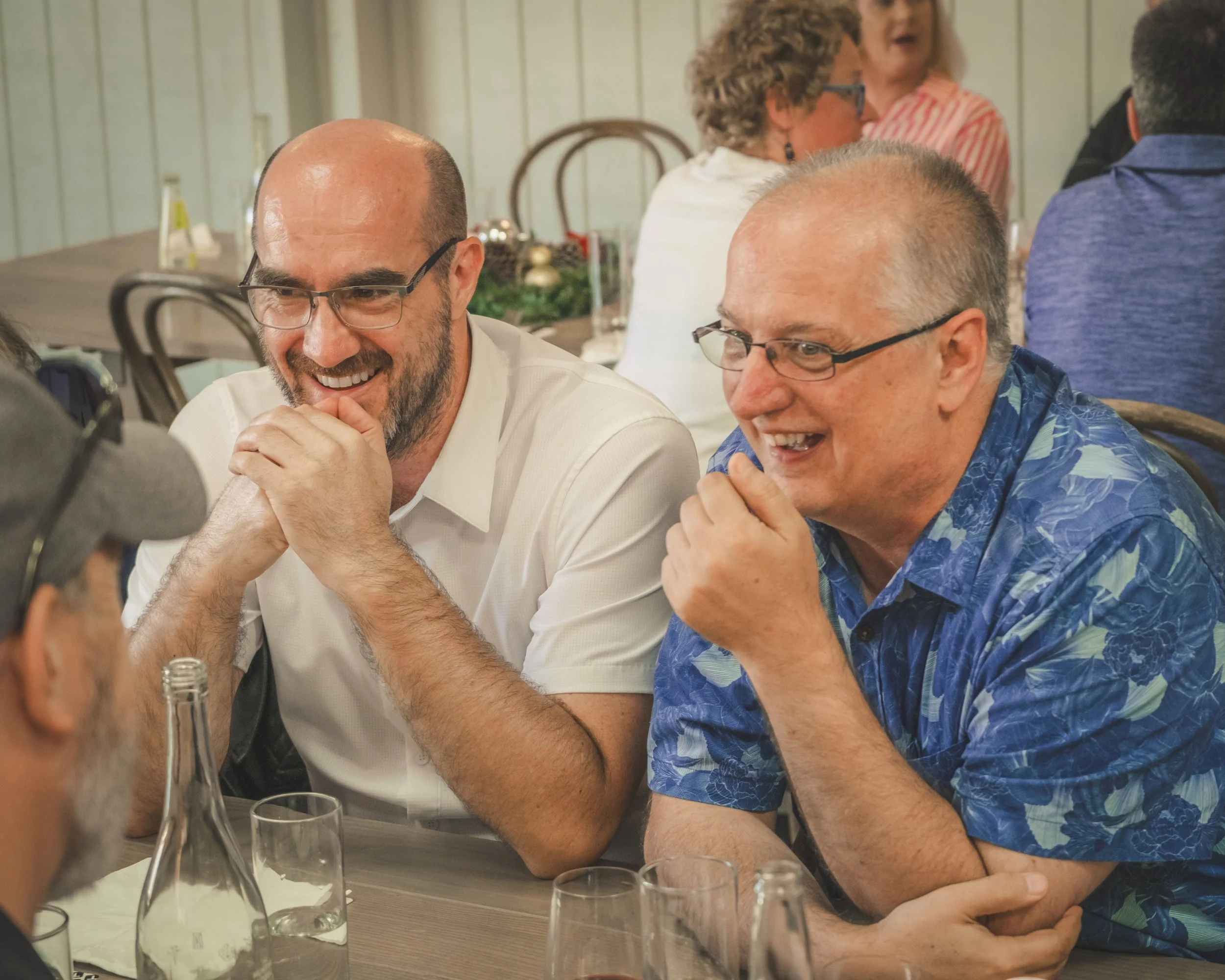 Two men with glasses and bald heads laughing and talking at a dinner table, surrounded by glasses and bottles, with other people in the background.