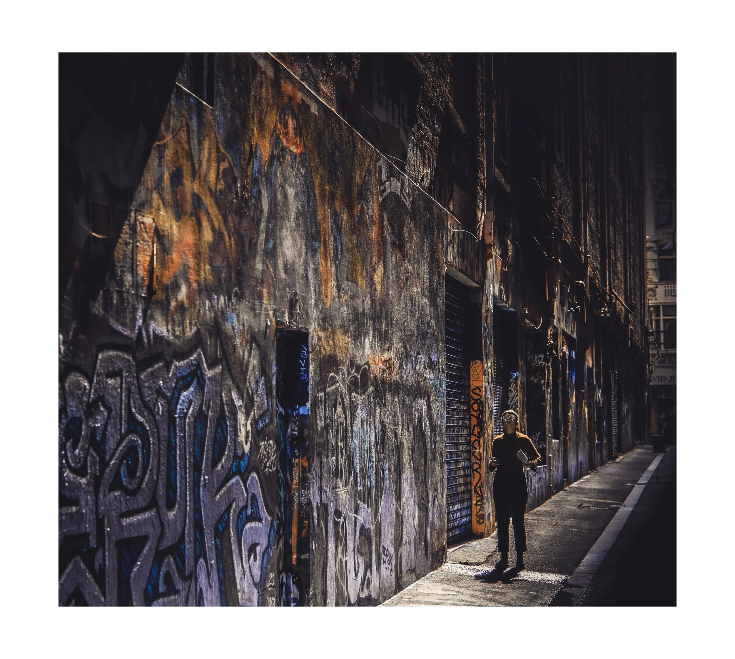 A person standing on a city sidewalk, next to a building covered in colourful graffiti and street art, with a look of wonder amongst the dark buildings in the background.