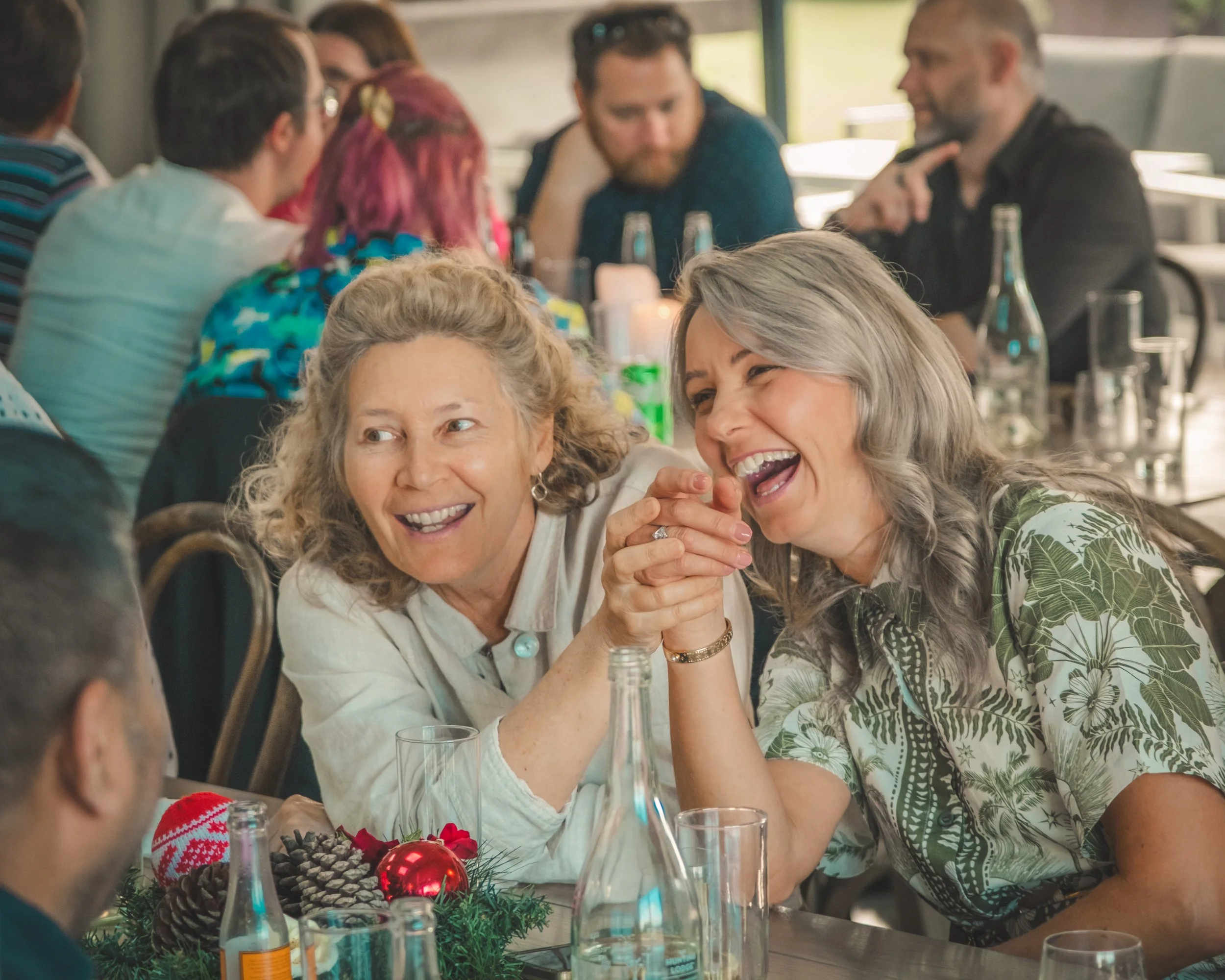 Two women are laughing and holding hands at a festive gathering with Christmas decorations on the table, surrounded by other people.
