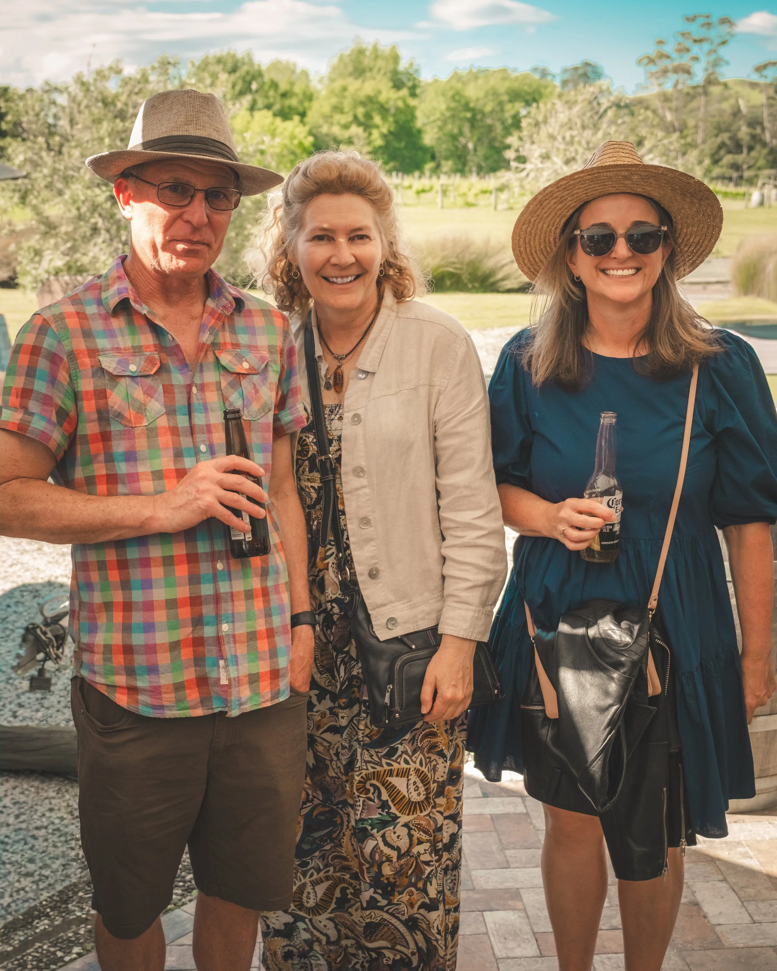 Three people standing outdoors in a park or garden area, smiling at the camera during daytime, with trees and blue sky in the background. The person on the left is a man wearing a straw hat, glasses, a plaid shirt, and holding a bottle of beer. The m