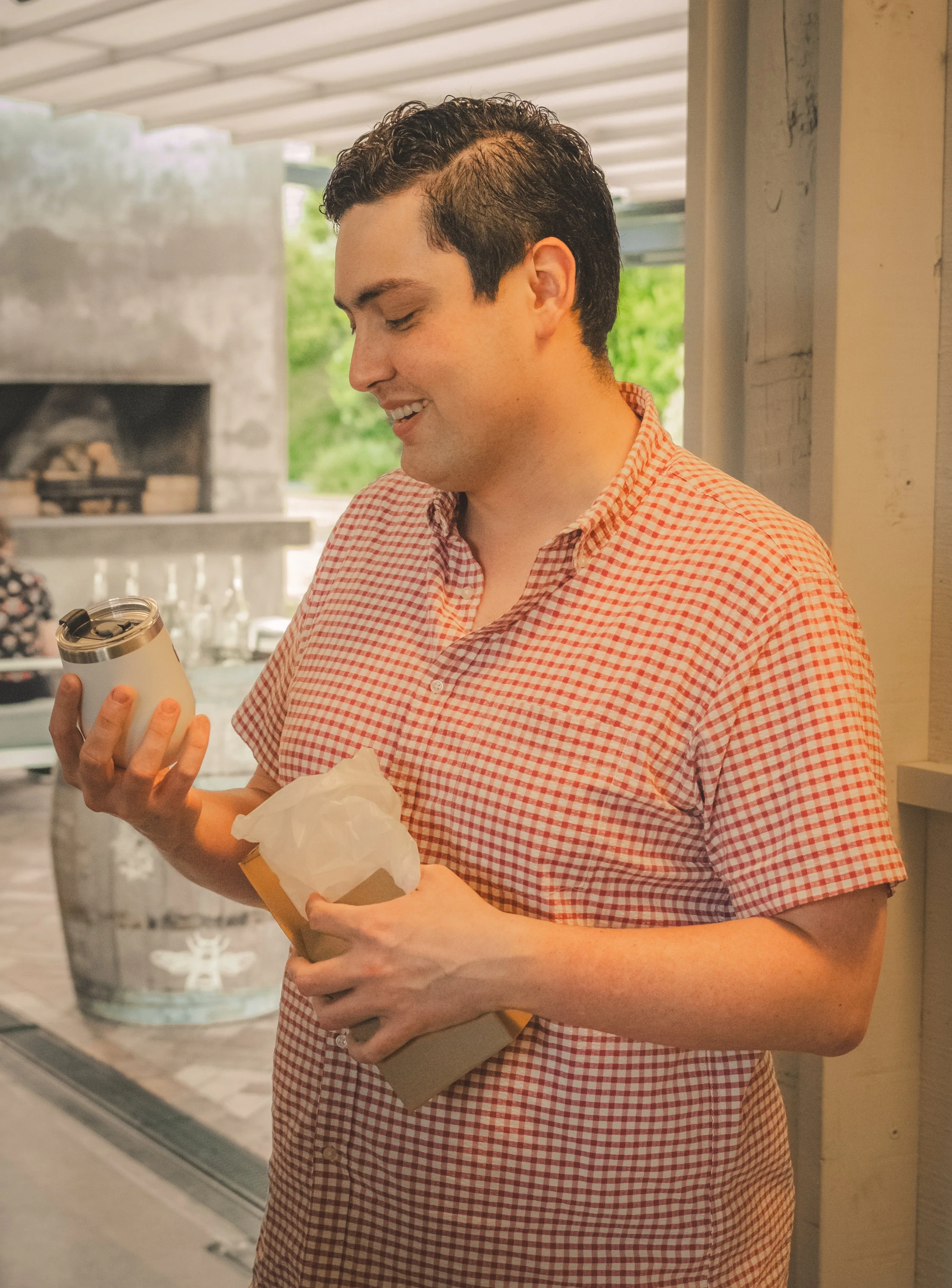 A man with dark, curly hair wearing a red and white checkered shirt, smiling while holding a white take-out coffee cup and a paper bag, standing outdoors near a brick oven or fireplace.