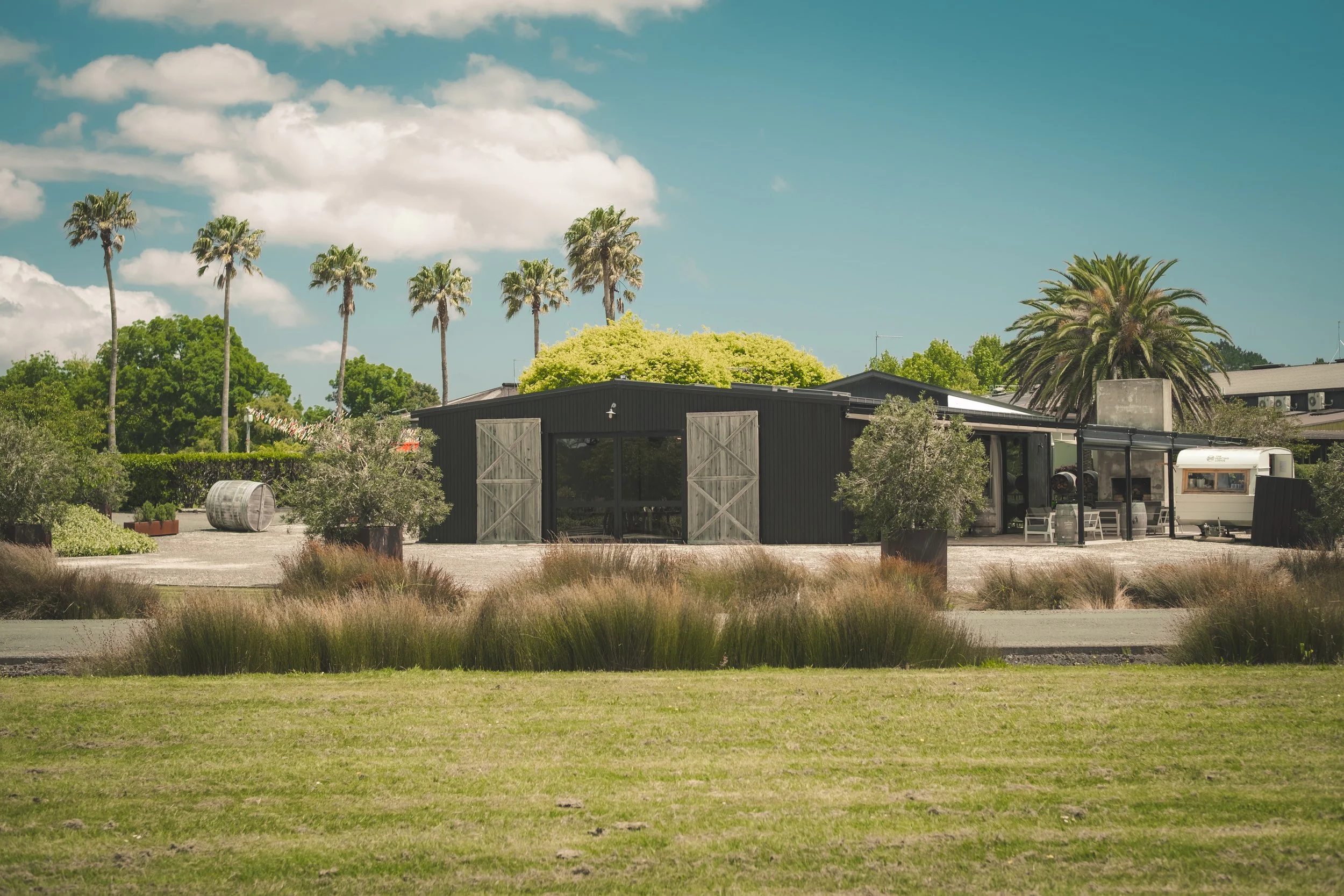 A clear sky with fluffy clouds over a rustic outdoor scene with palm trees, a black building with barn doors, potted plants, and outdoor seating. There are also large barrels and a trailer visible.
