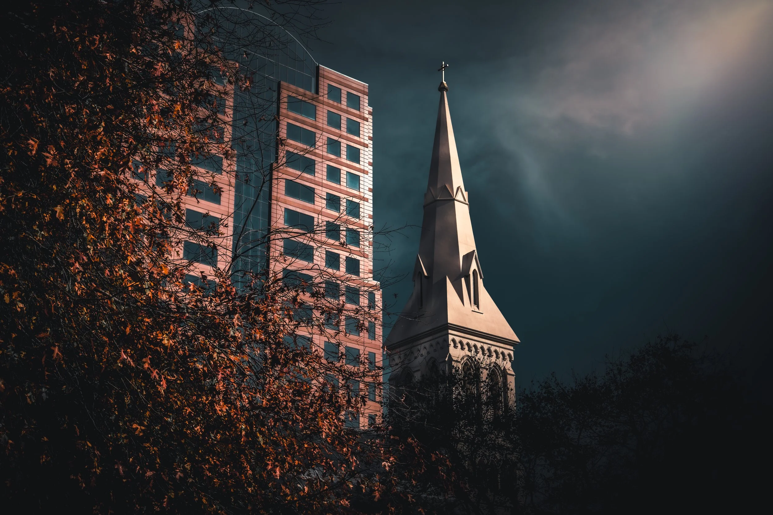 A tall church steeple with a cross on top, next to a modern glass building, with trees in the foreground and a dark, cloudy sky in the background.