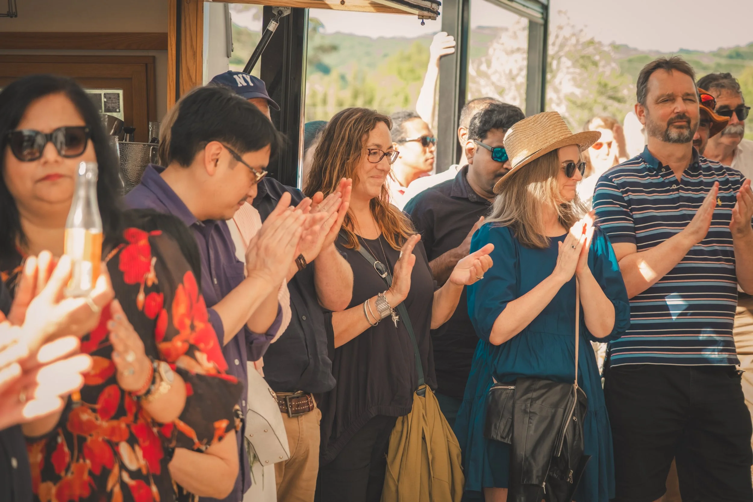 People standing close together with hands clasped or raised in prayer, some with eyes closed, inside a building with large windows showing an outdoor landscape.