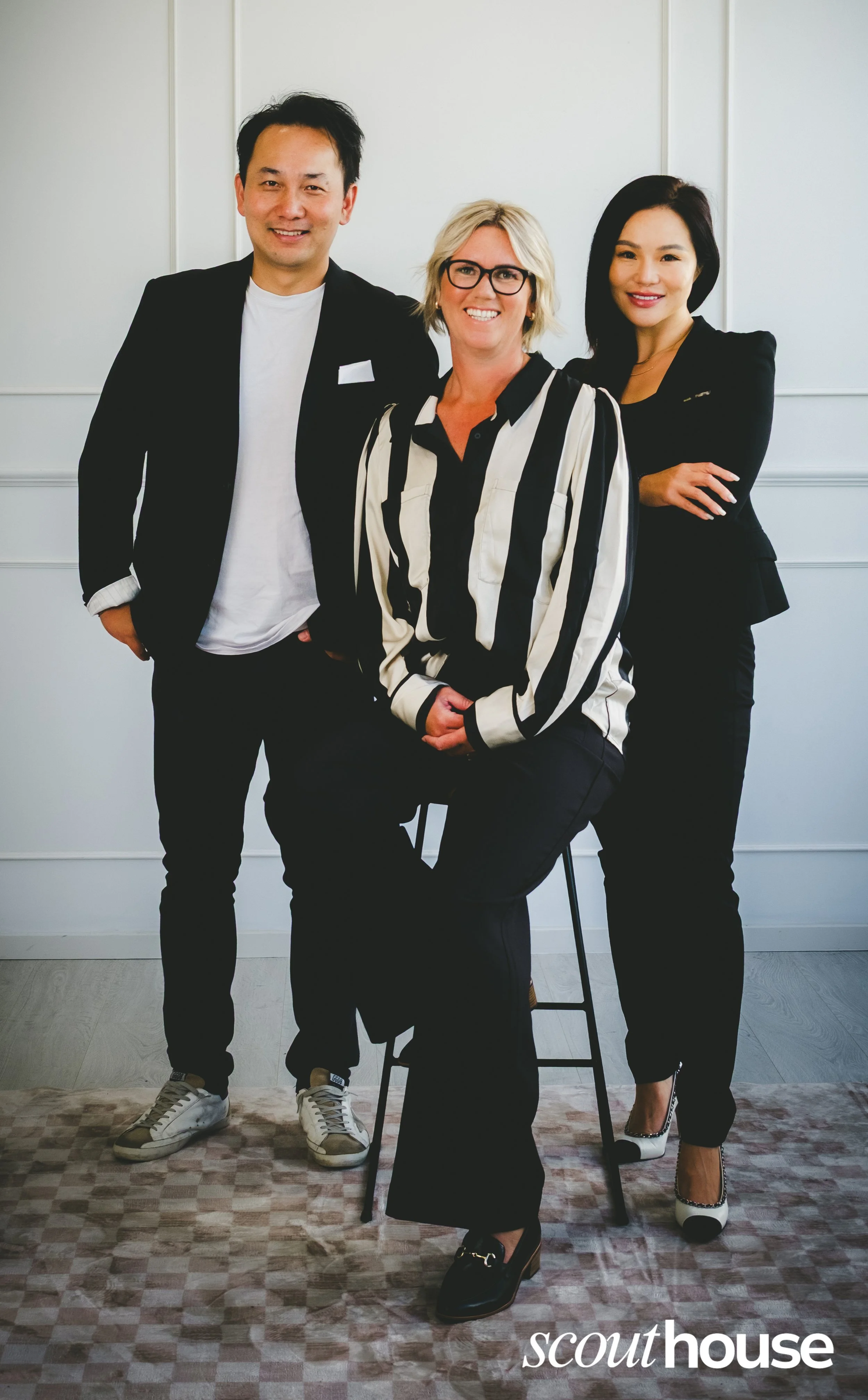 Three people posing for a photo in a professional setting, with two women and one man. The woman in the middle has blonde hair, glasses, and is wearing a black and white striped blouse. The man on the left has short dark hair and is wearing a black b