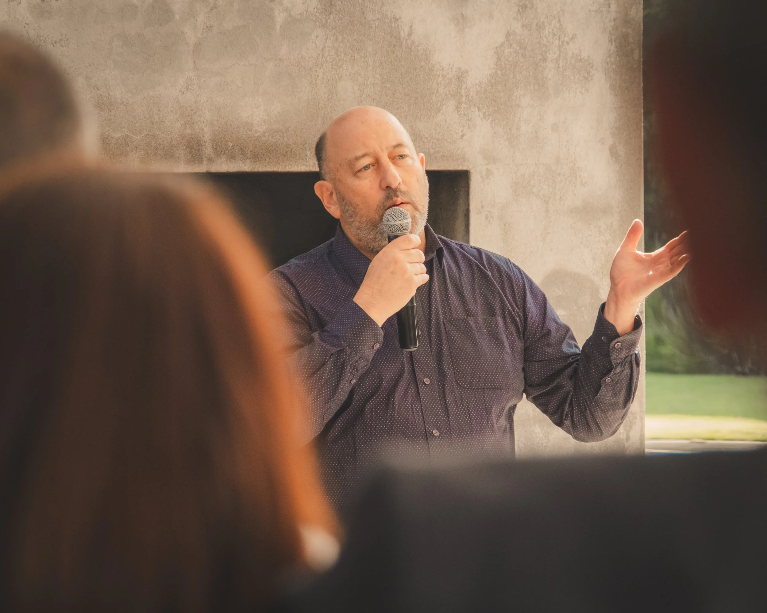 A man with a beard and bald head speaking into a microphone with a hand gesture, captured in an indoor setting with a concrete wall background.