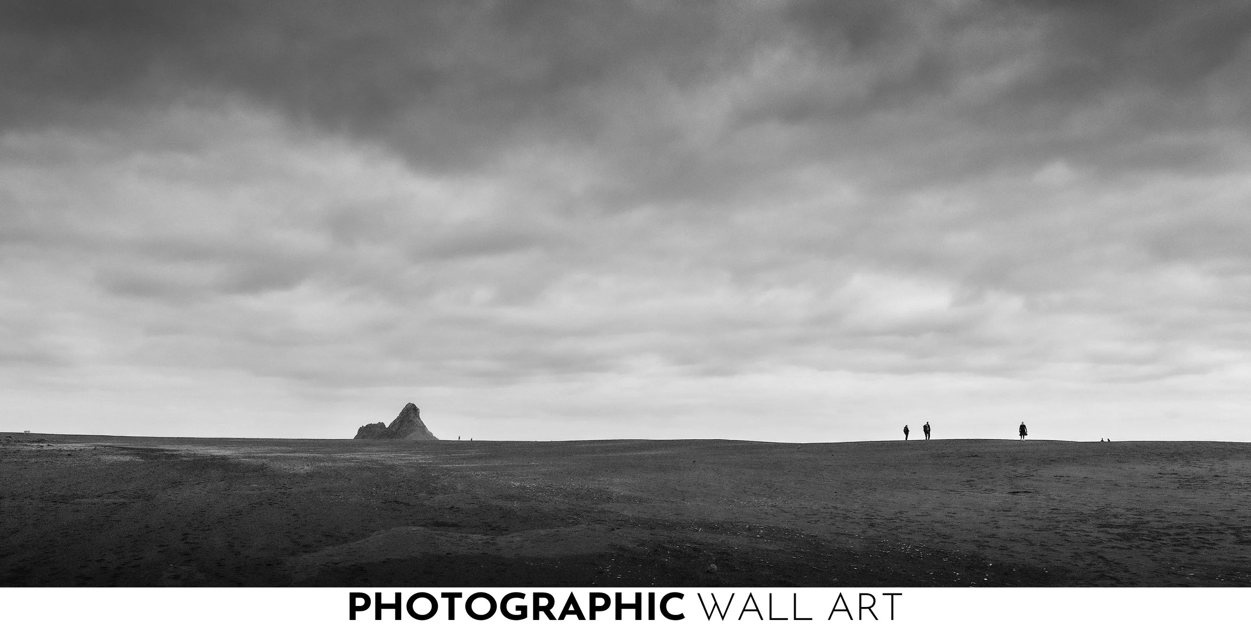 An artistic black and white photo of a coastal landscape with dark sky and clouds, a mountain in the distance, and three small figures walking along the shore. The image evokes an emotional reaction to the loneliness of the location.