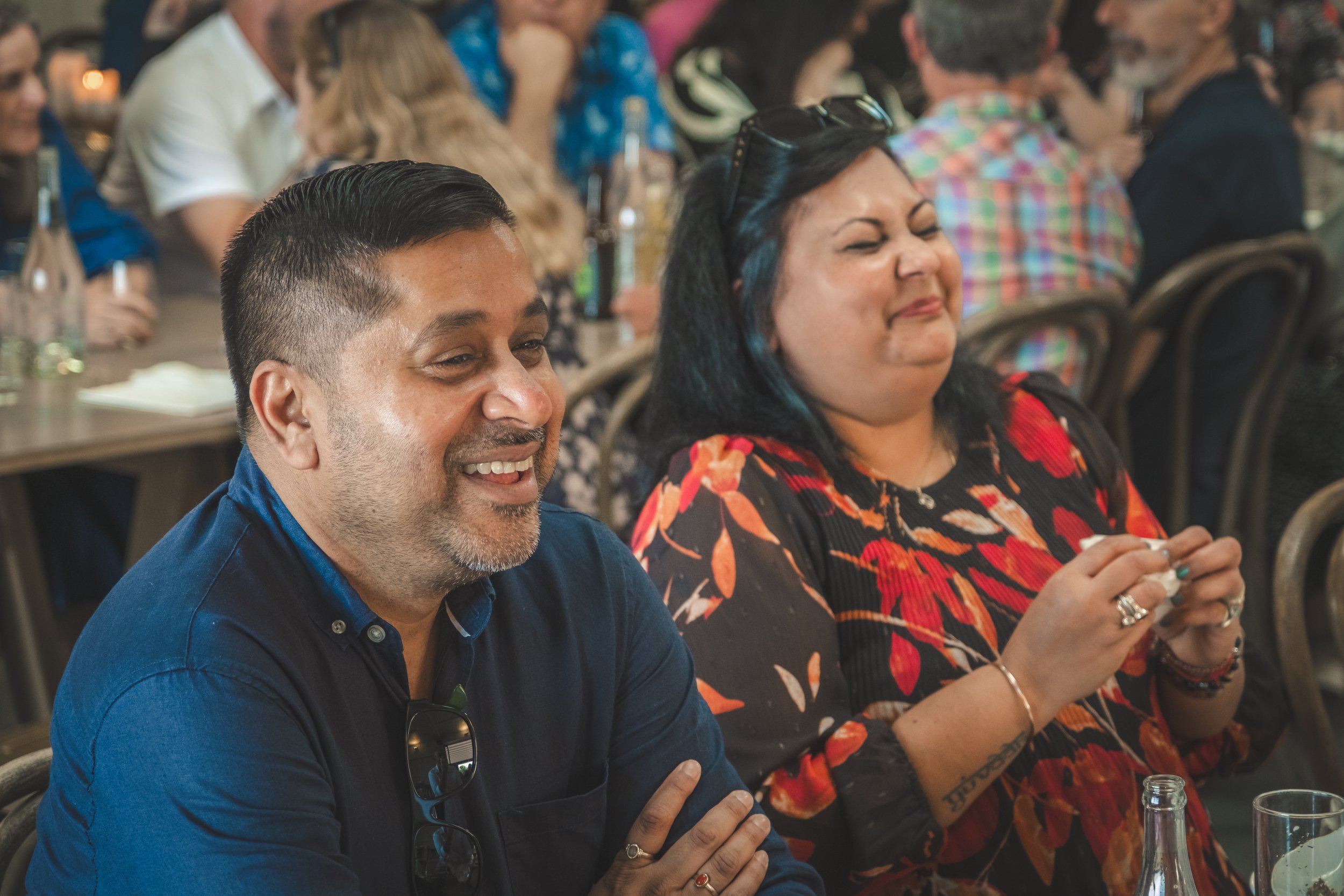 A group of diverse people seated at a conference table, some smiling and engaging, with bottles and glasses of drinks on the table, in a warm, lively setting.