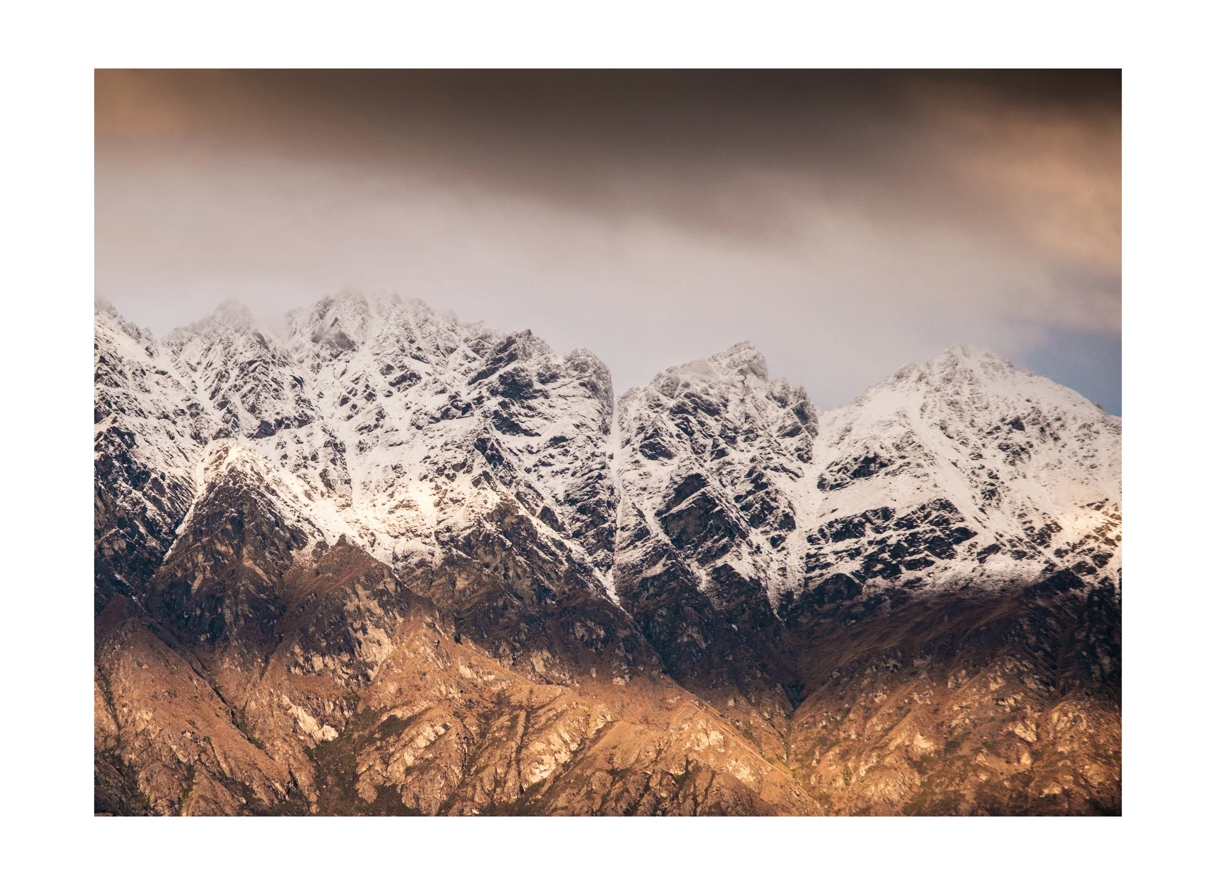 Snow-covered Remarkables Mountain Range with rocky slopes beneath cloudy sky at dusk.