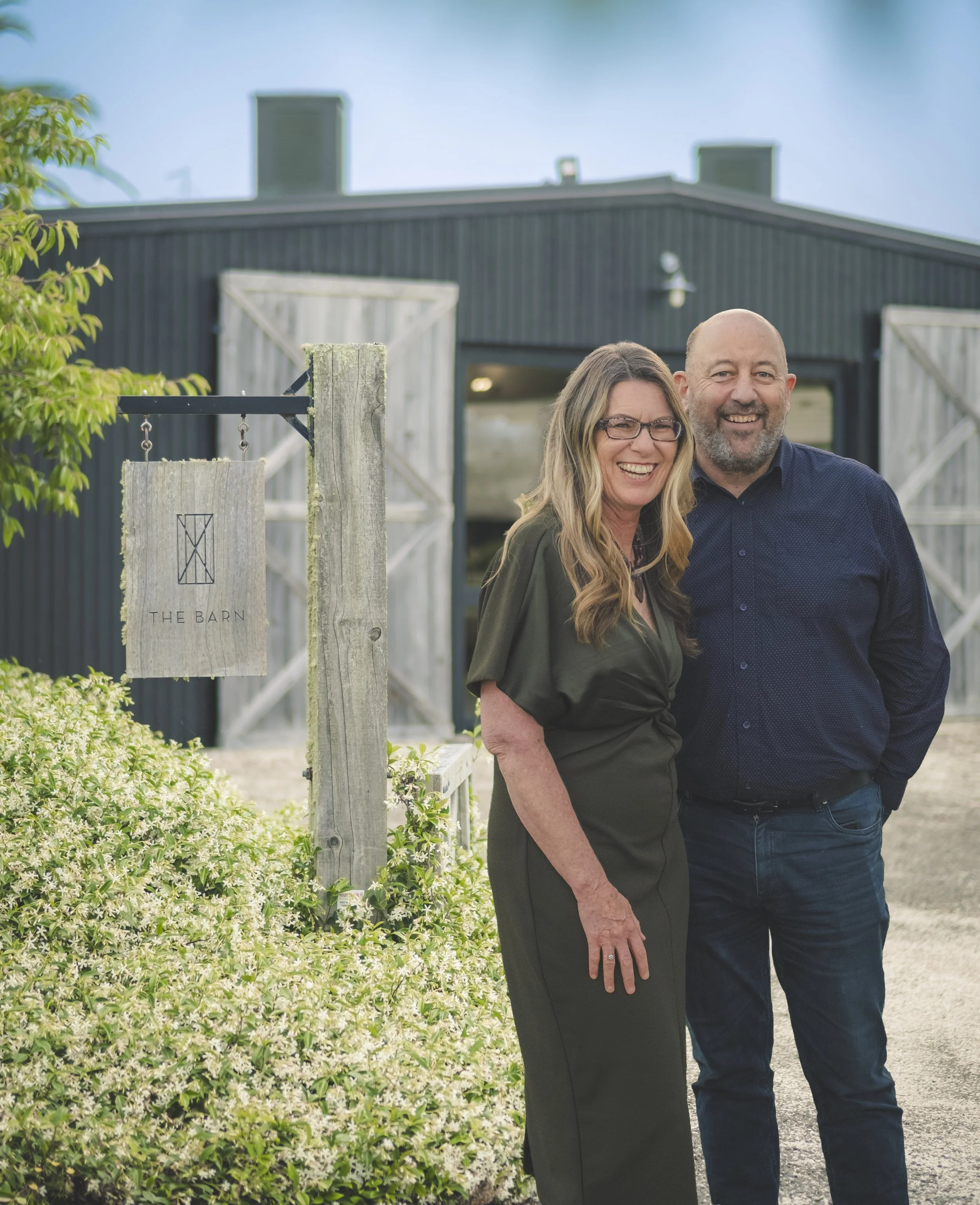 A smiling woman with glasses and a man with a beard standing outside in front of a barn and a garden with green bushes and white flowers.
