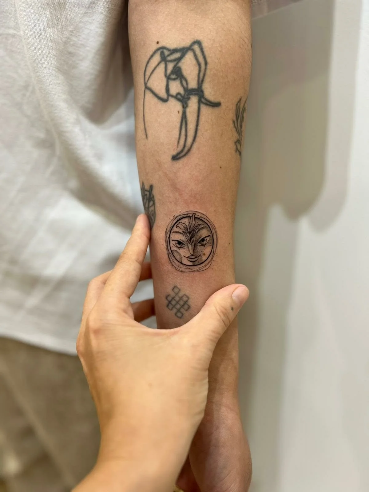 A person's arm with three tattoos: a line drawing of a person holding a camera, a circular face of a feline with detailed features, and a geometric pattern below the face.