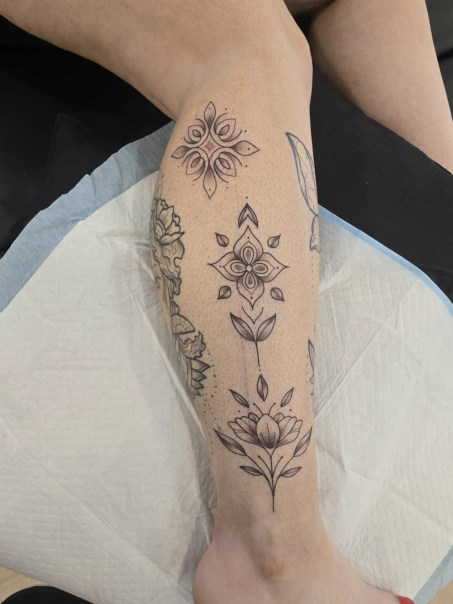 A person's lower leg with a fresh beautiful fine line black ink floral tattoo design, featuring geometric and organic elements, spread vertically along the calf. Heart and home tattoo studio moonee ponds 