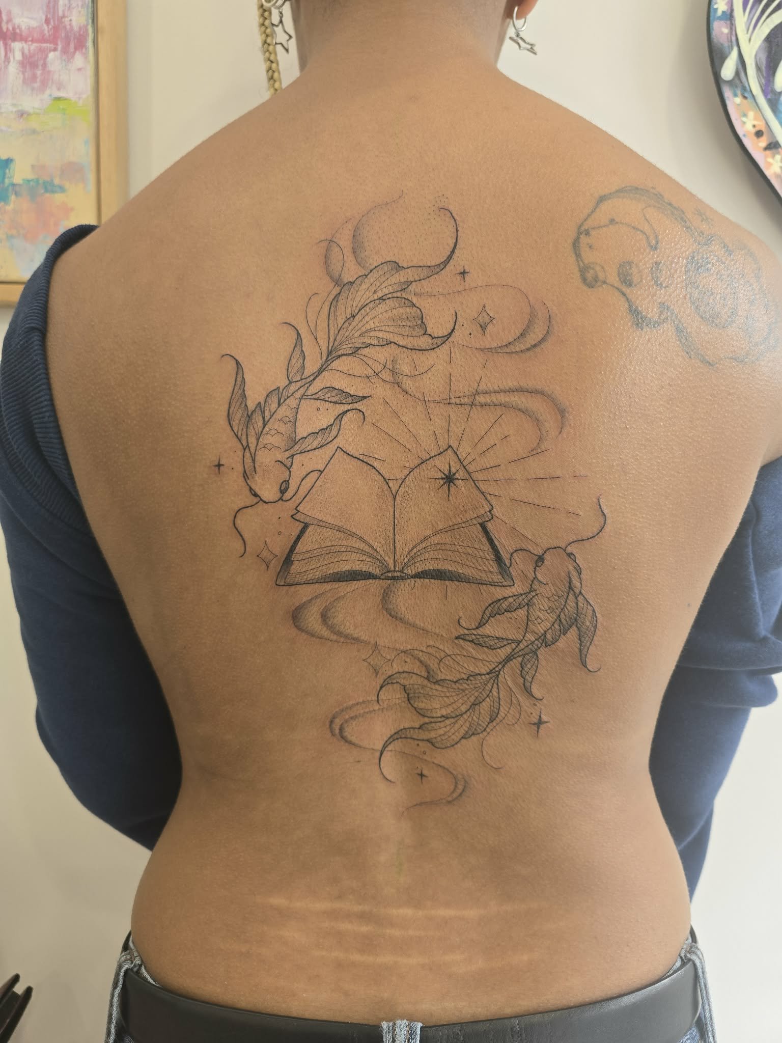 Fine line Tattoo on a person's back featuring a fish, an open book with rays of light, stars, moons, and swirling clouds or smoke. Melbourne mooned ponds tattoo, best tattoo Melbourne 