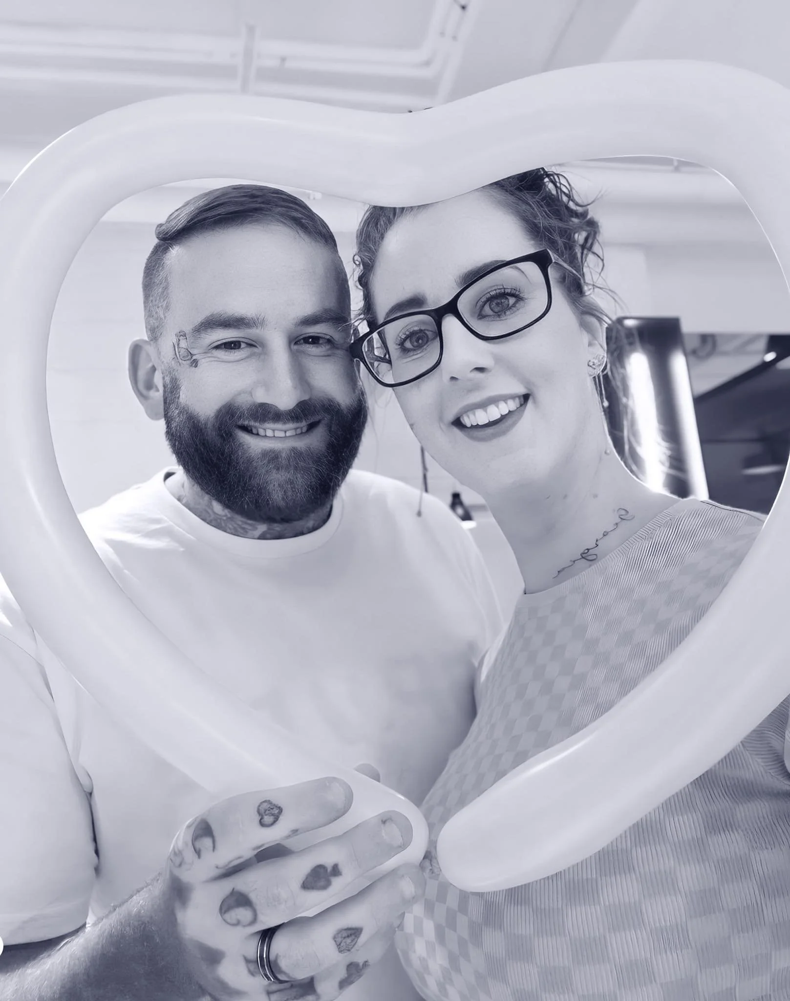 A man and woman take a selfie behind a large heart-shaped frame, smiling and looking at the camera.