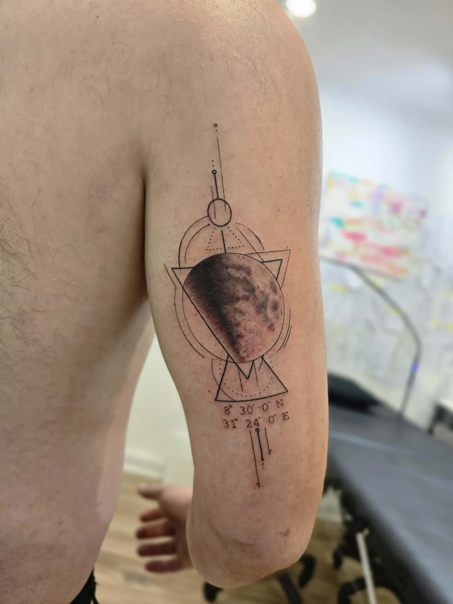 Tattoo of the moon with geometric shapes and coordinates on person's upper arm.
